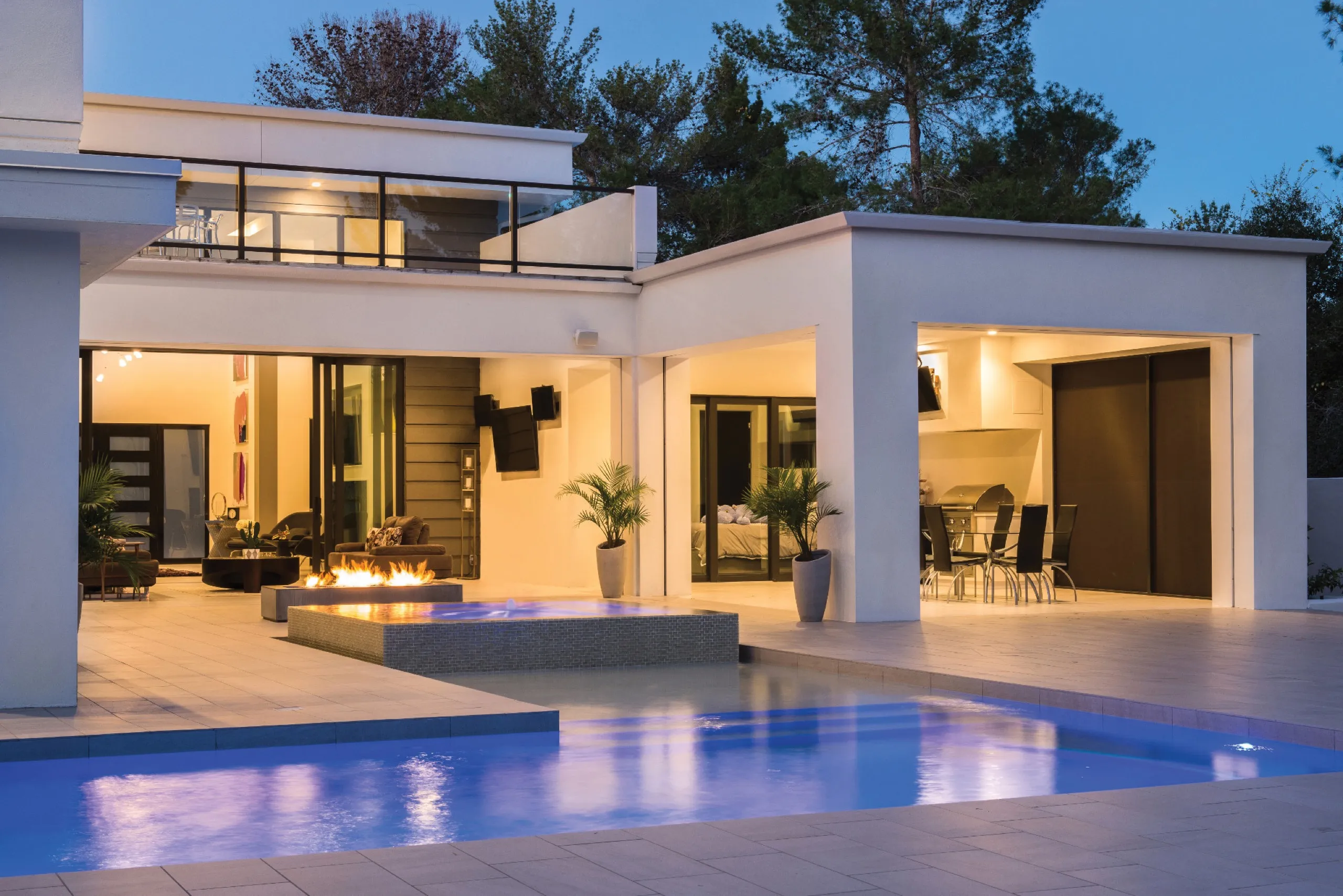 Modern backyard at dusk with a blue-lit pool, fire pit, outdoor dining patio, and wall-mounted speakers.