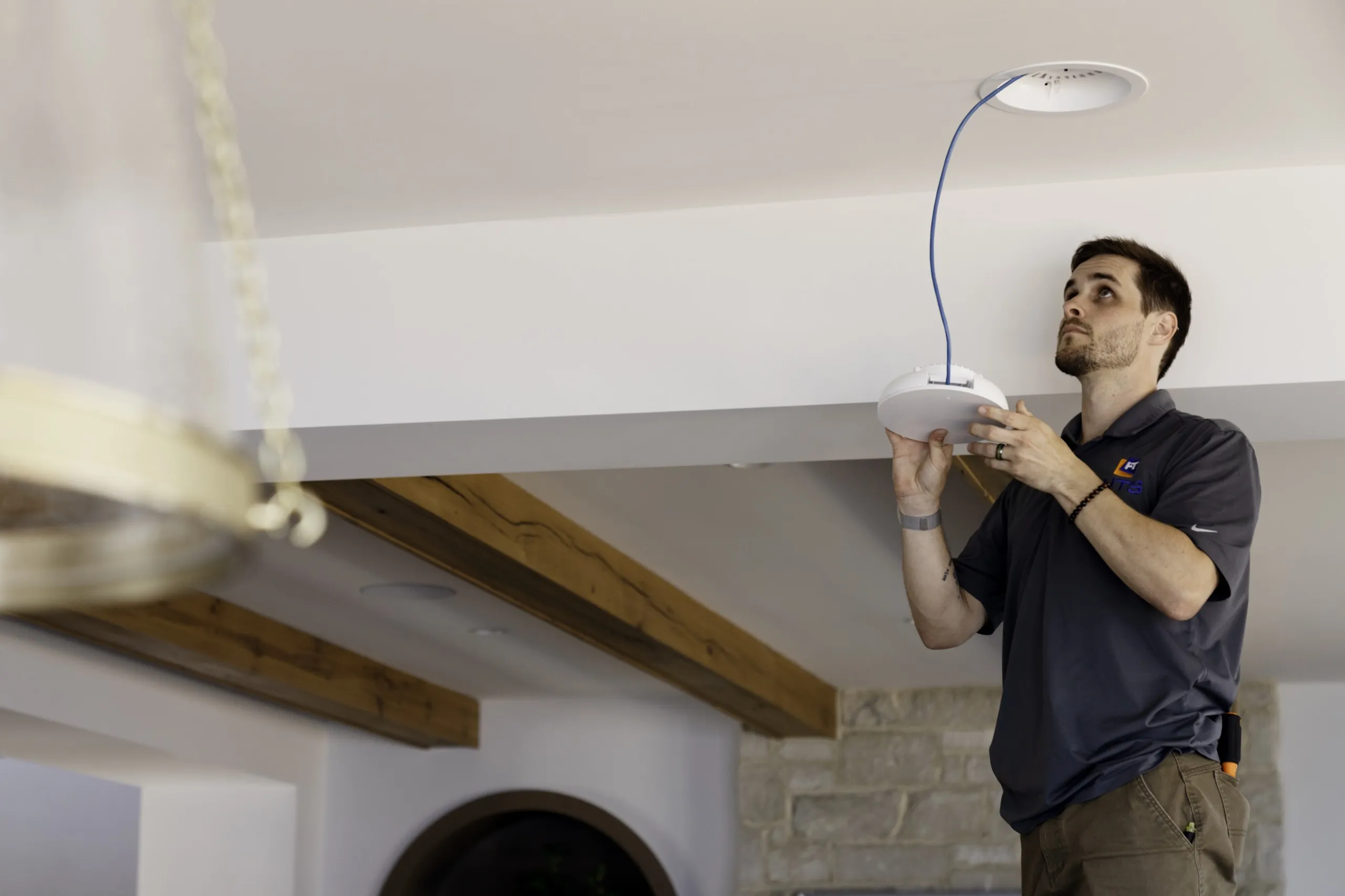 Installer lifting a white ceiling Wi-Fi access point toward a blue network cable in a room with wood beams.