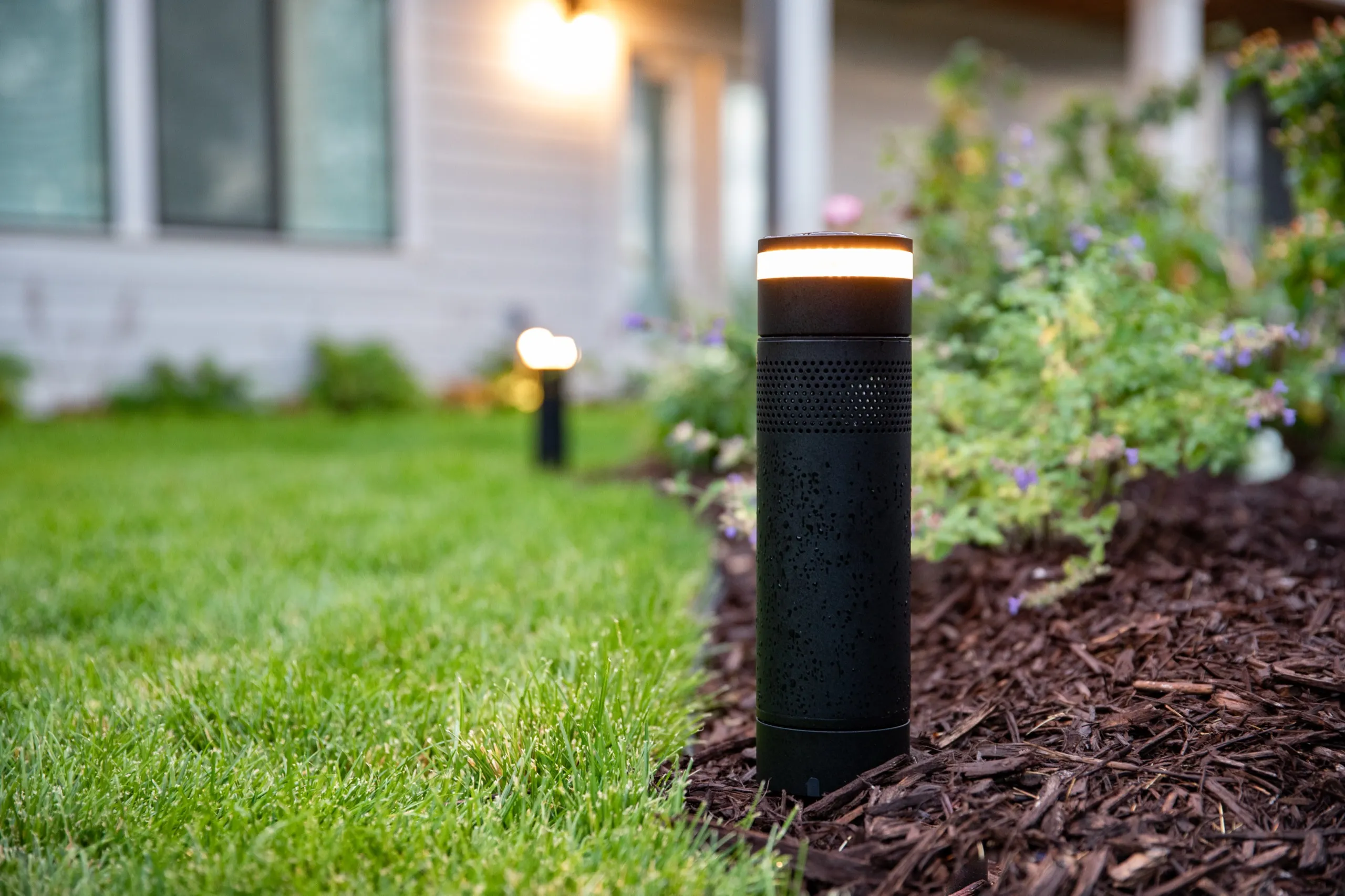 Black outdoor landscape bollard speaker-light glowing on a mulched garden bed next to a green lawn.