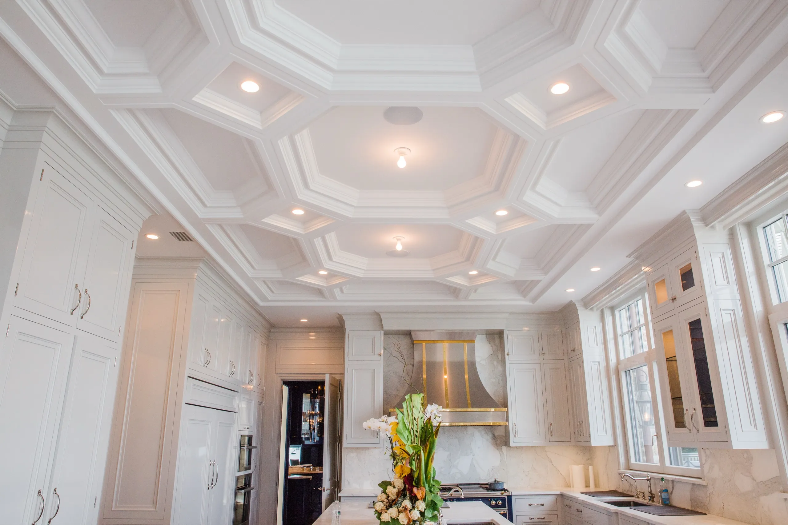 Luxury white kitchen with a geometric coffered ceiling, recessed lighting, brass range hood, and marble surfaces.