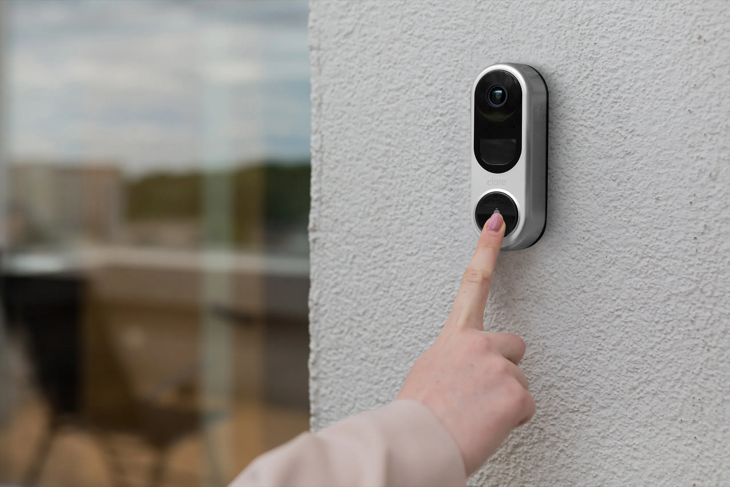 Hand pressing a silver video doorbell with dual cameras on a white exterior wall.