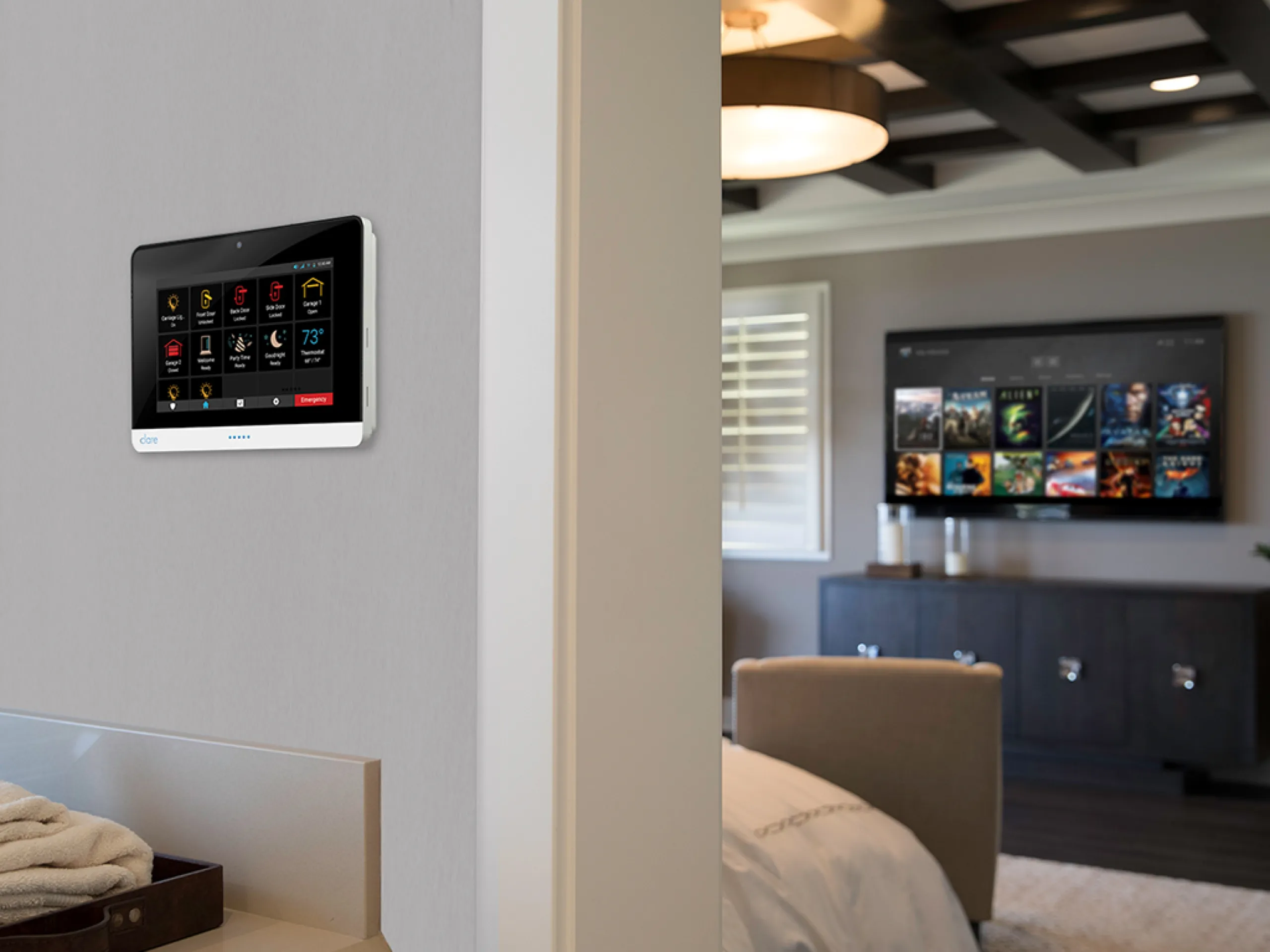 Wall-mounted home security touchscreen panel beside a bedroom with a large TV.
