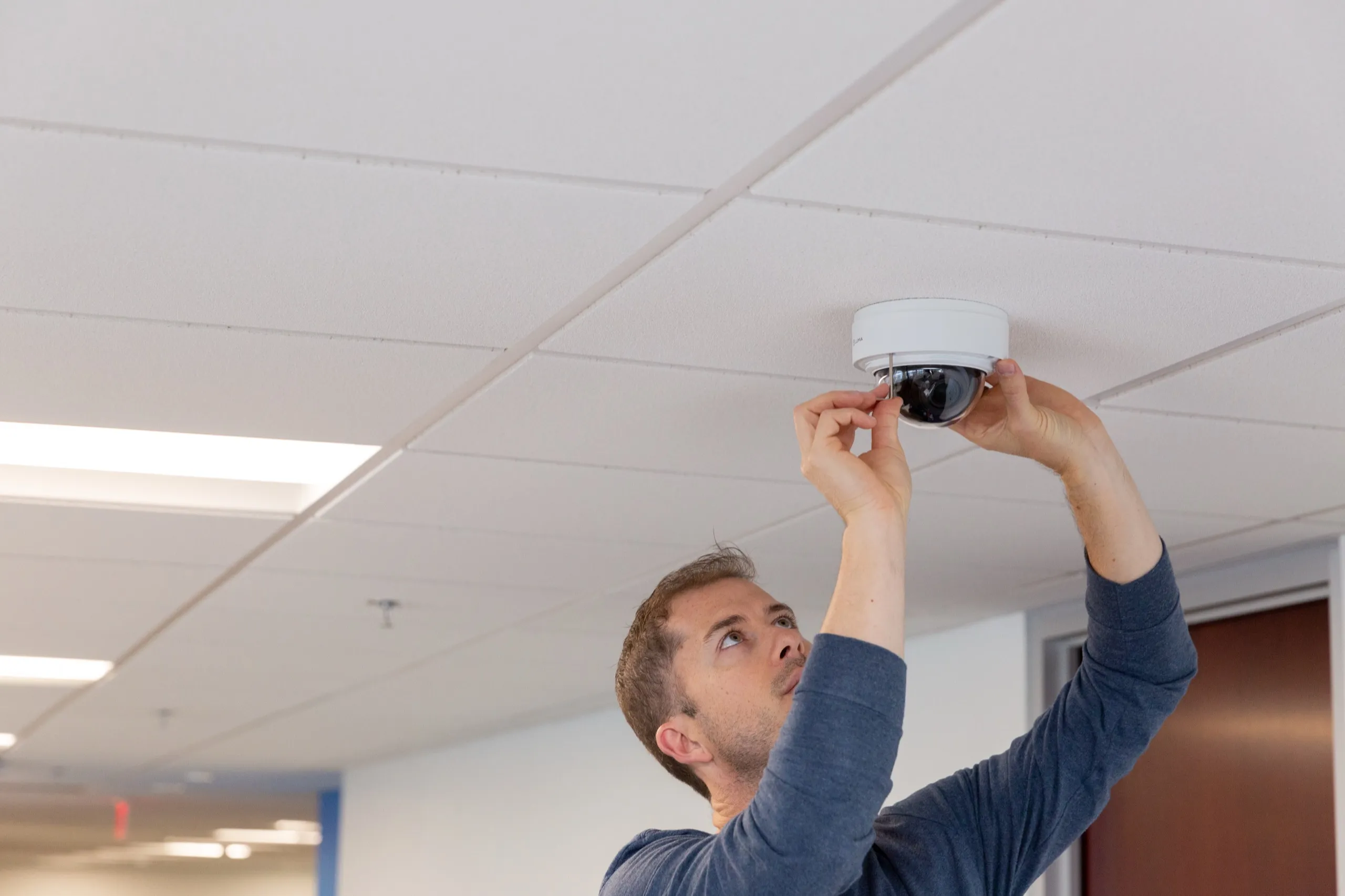Technician mounting a white dome security camera on a drop ceiling in a commercial corridor.