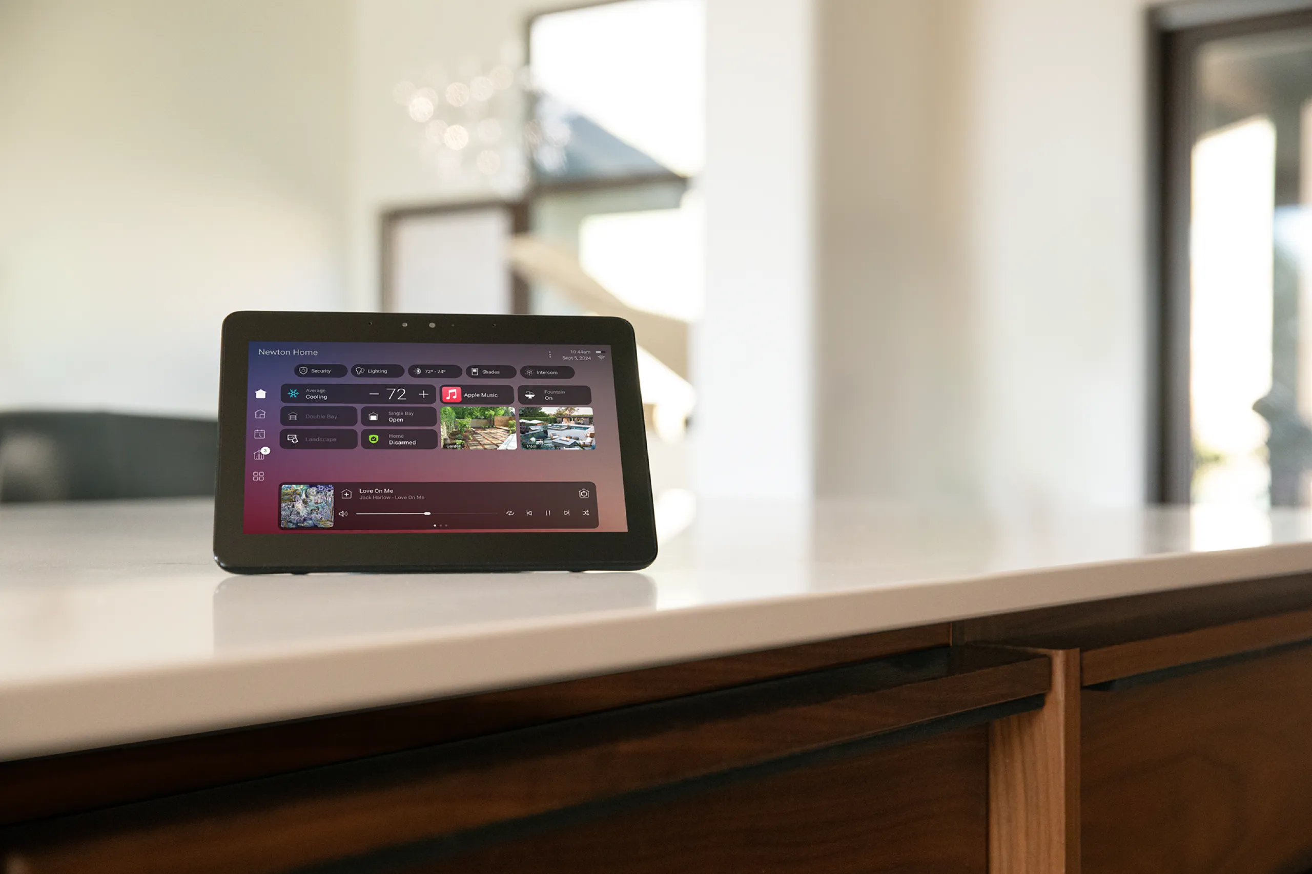 Black smart-home tablet on a kitchen counter showing climate, cameras, and music controls.