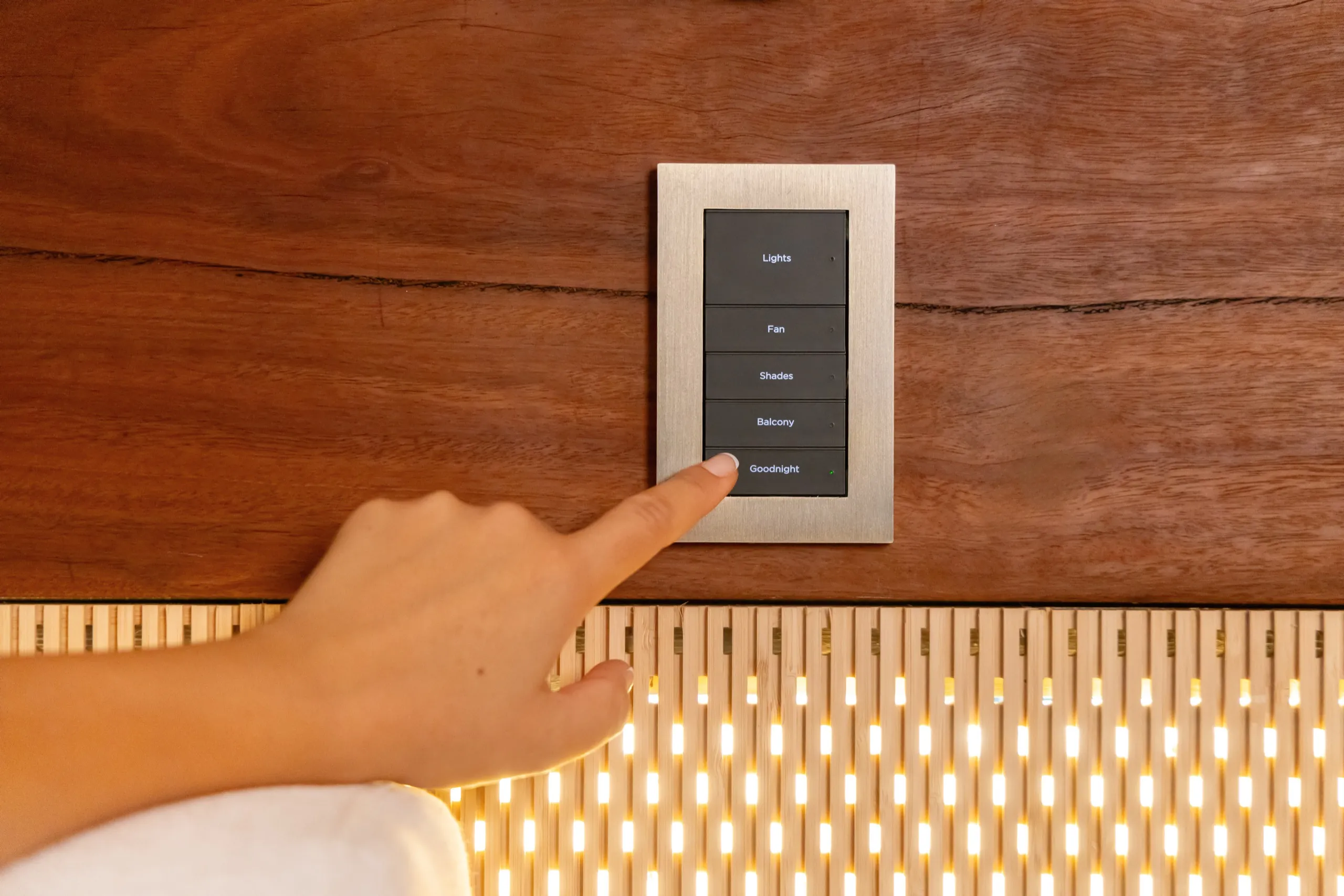 Finger pressing the Goodnight button on a silver wall keypad mounted on dark wood paneling.
