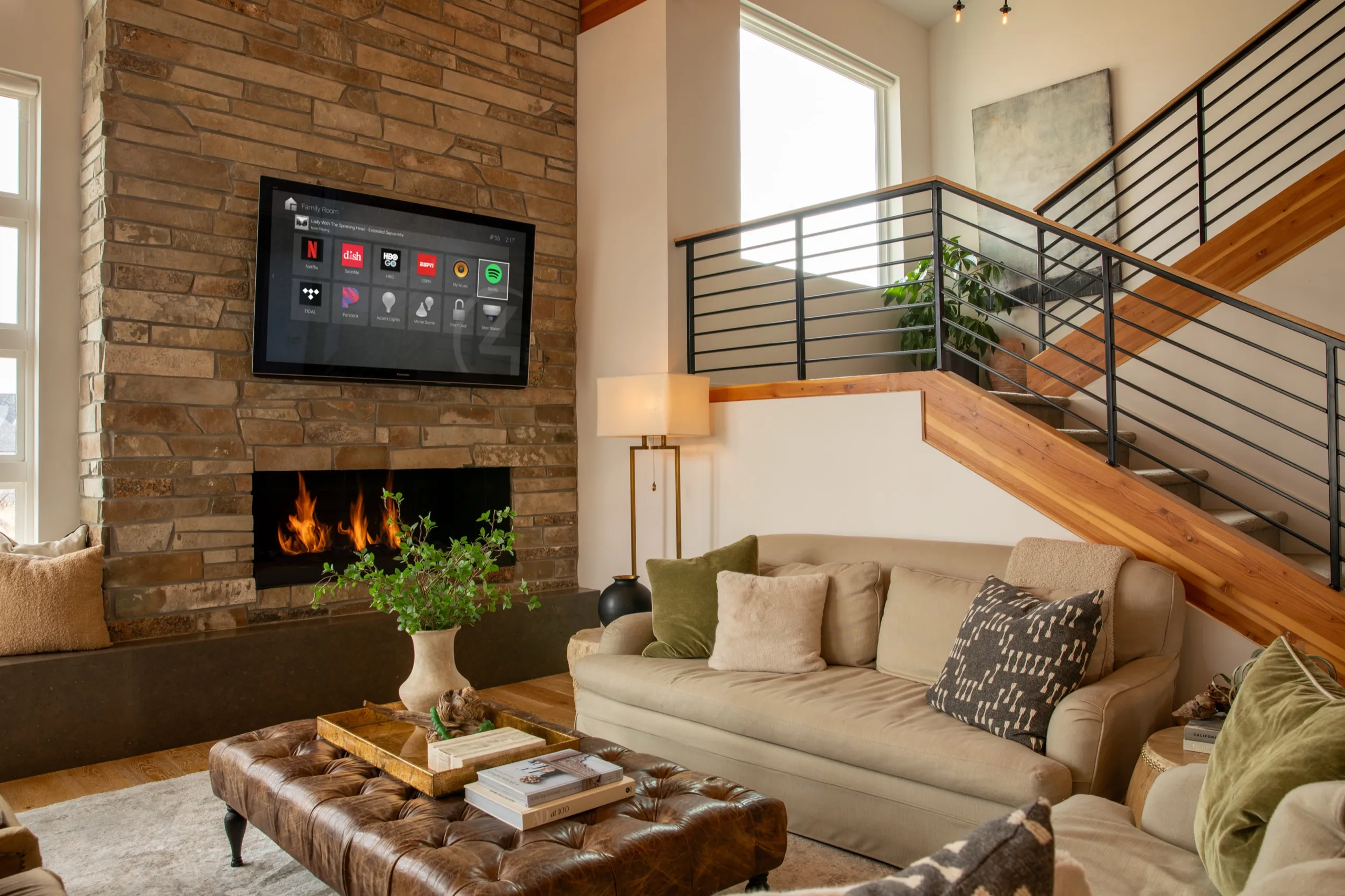 Modern living room with stone fireplace, wall TV showing smart-home apps, sofa seating, and wood staircase.