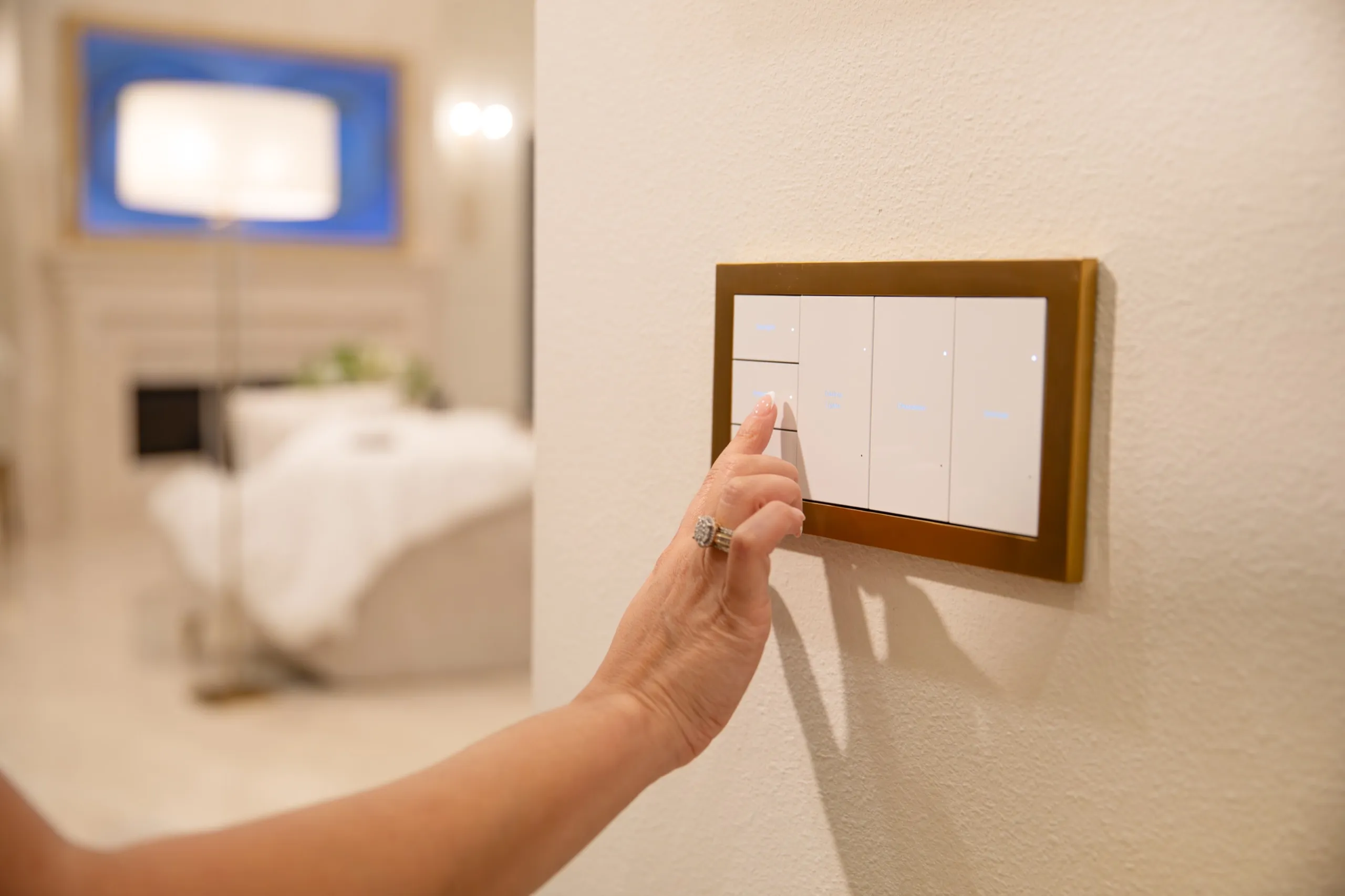 Hand pressing a gold-framed multi-button light keypad on a wall with a room blurred in the background.