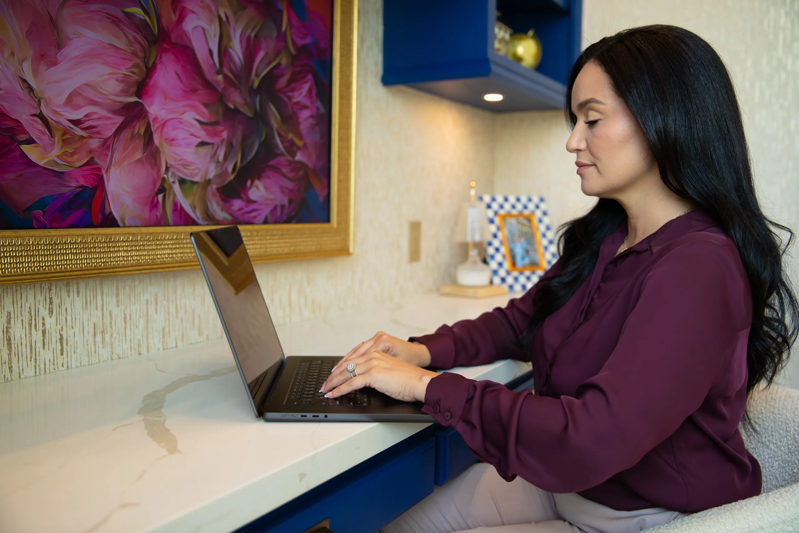 Woman working on a laptop at a modern desk under a large pink floral painting and blue cabinetry.