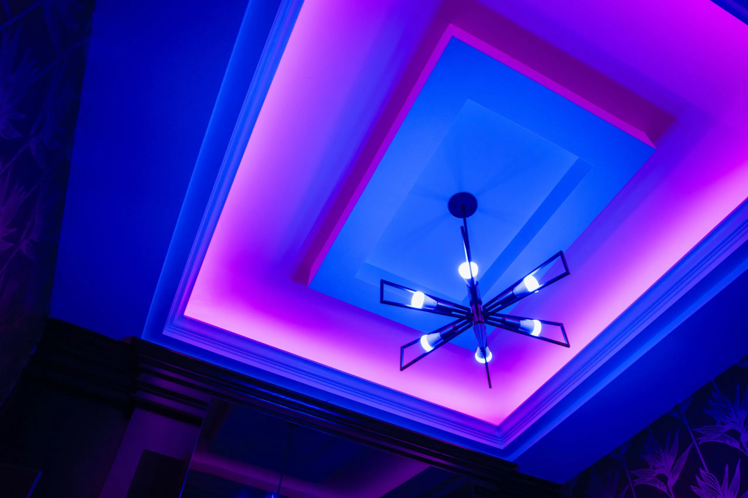 Multi-level coffered ceiling washed in colored cove lighting around a modern multi-arm chandelier.