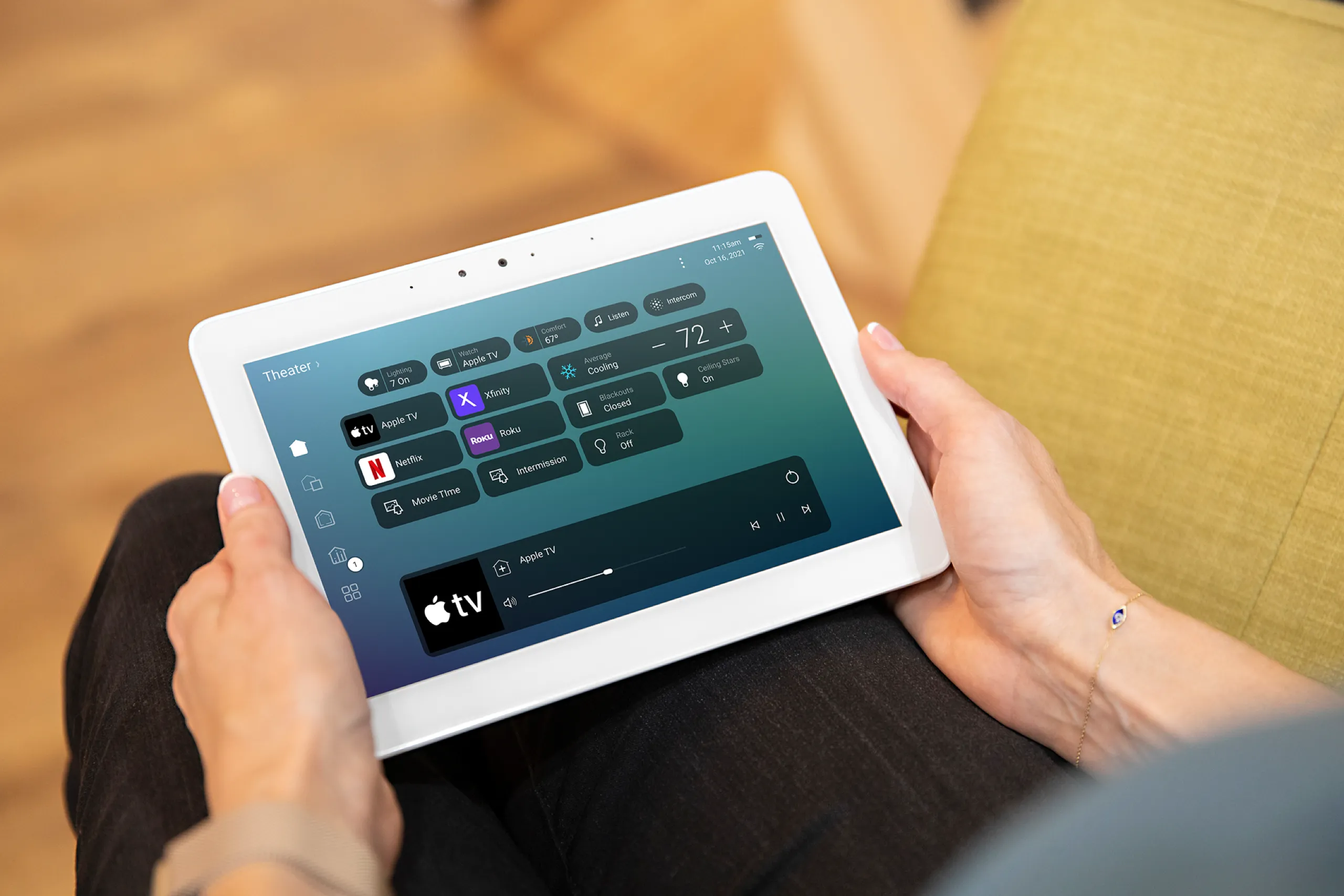 Hands holding a white smart-home tablet open to a theater control screen with streaming apps and volume for Apple TV.