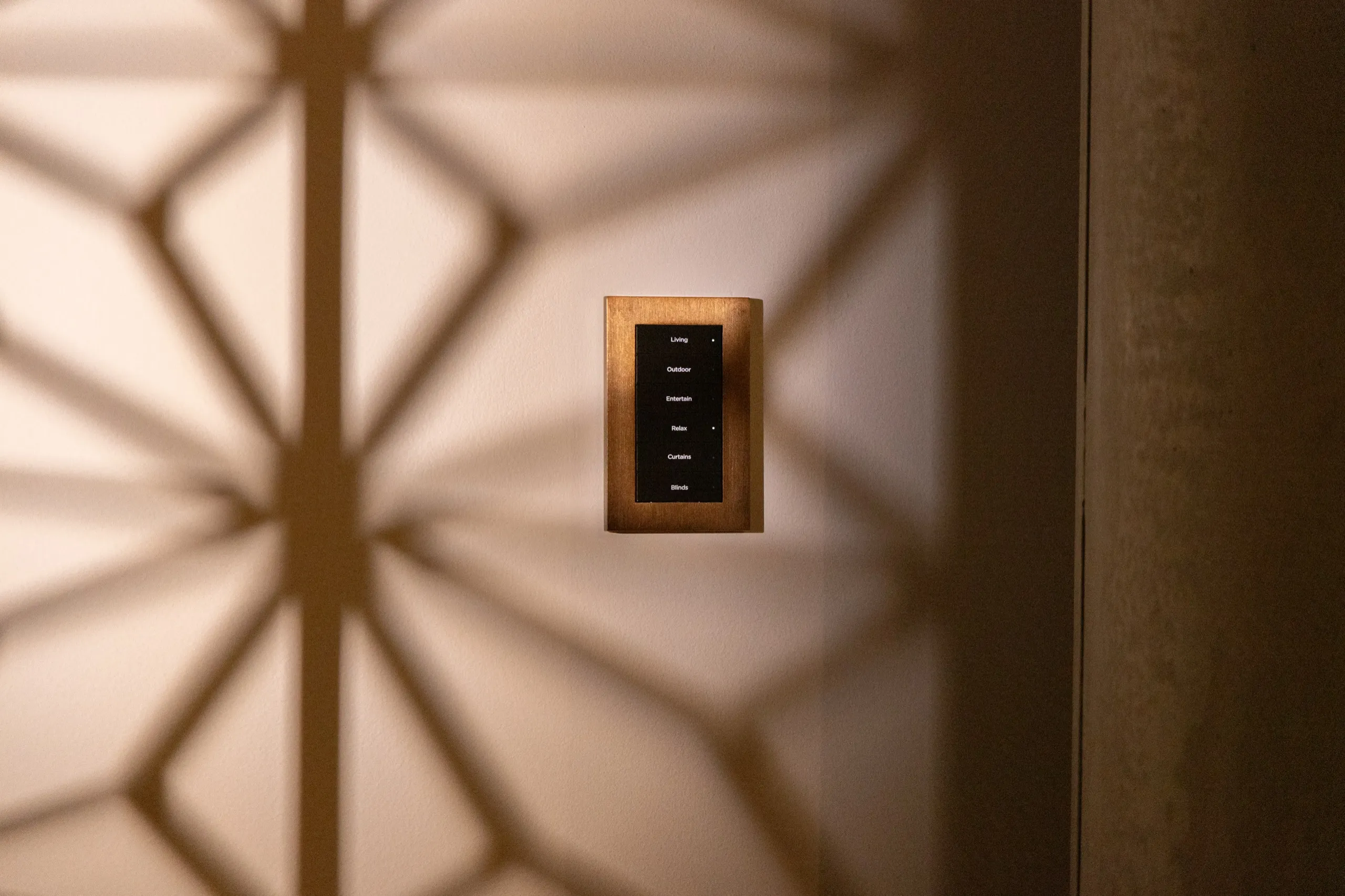 Slim wood-framed wall keypad with a black face on a cream wall under geometric lattice shadows.