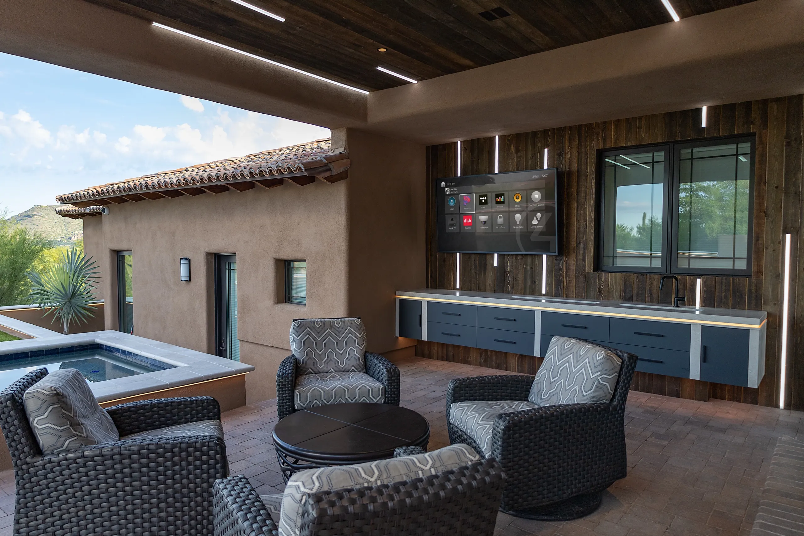 Covered patio with wicker chairs, outdoor kitchen bar, and wall TV showing smart-home controls near a pool.