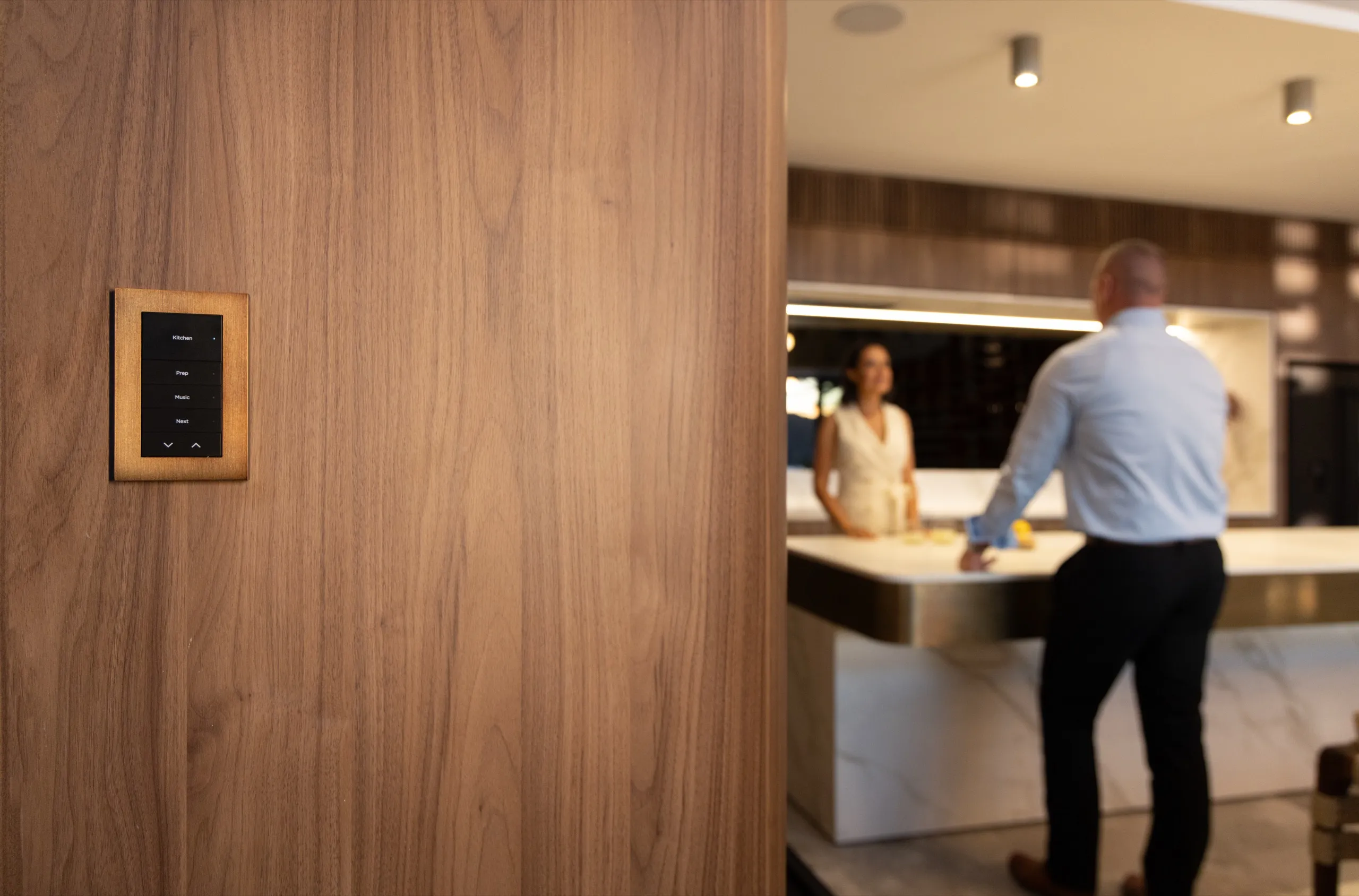 Wood-framed wall keypad in focus beside an open kitchen where a couple stands at a marble island.
