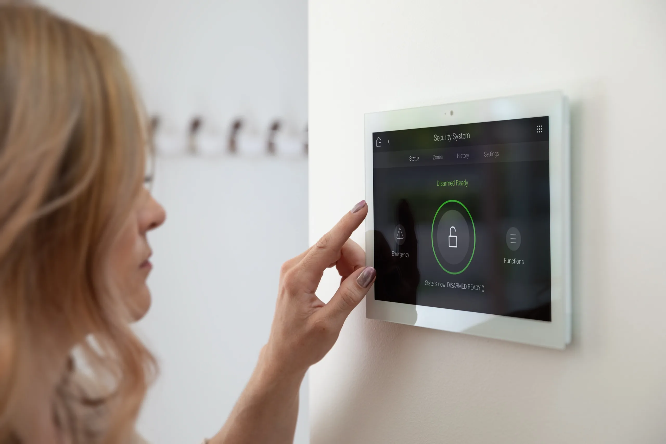 Woman touching a white wall touchscreen showing a security system disarmed-ready status.