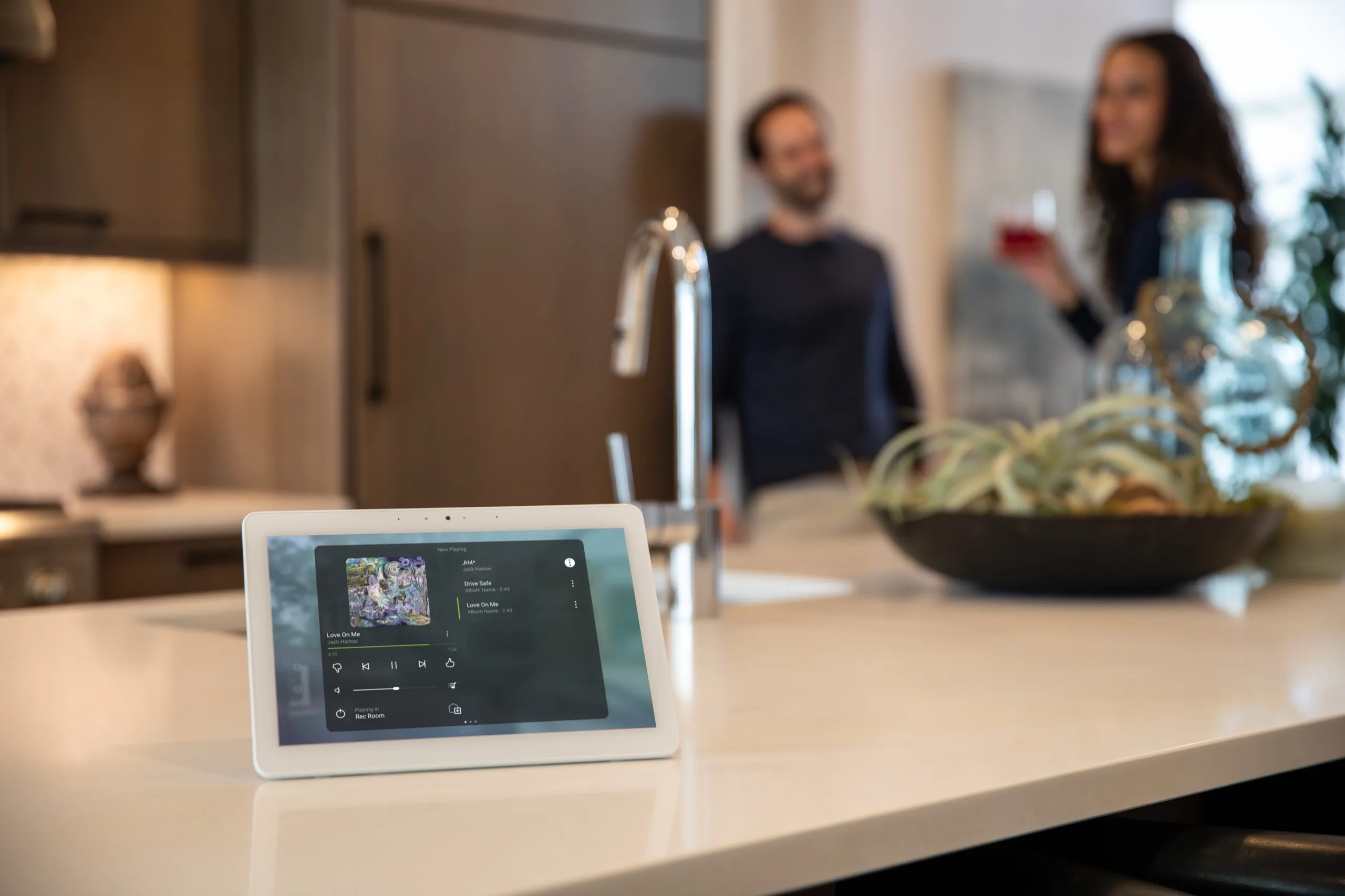 White smart-home tablet on a kitchen island playing music while a couple stands at the sink behind.