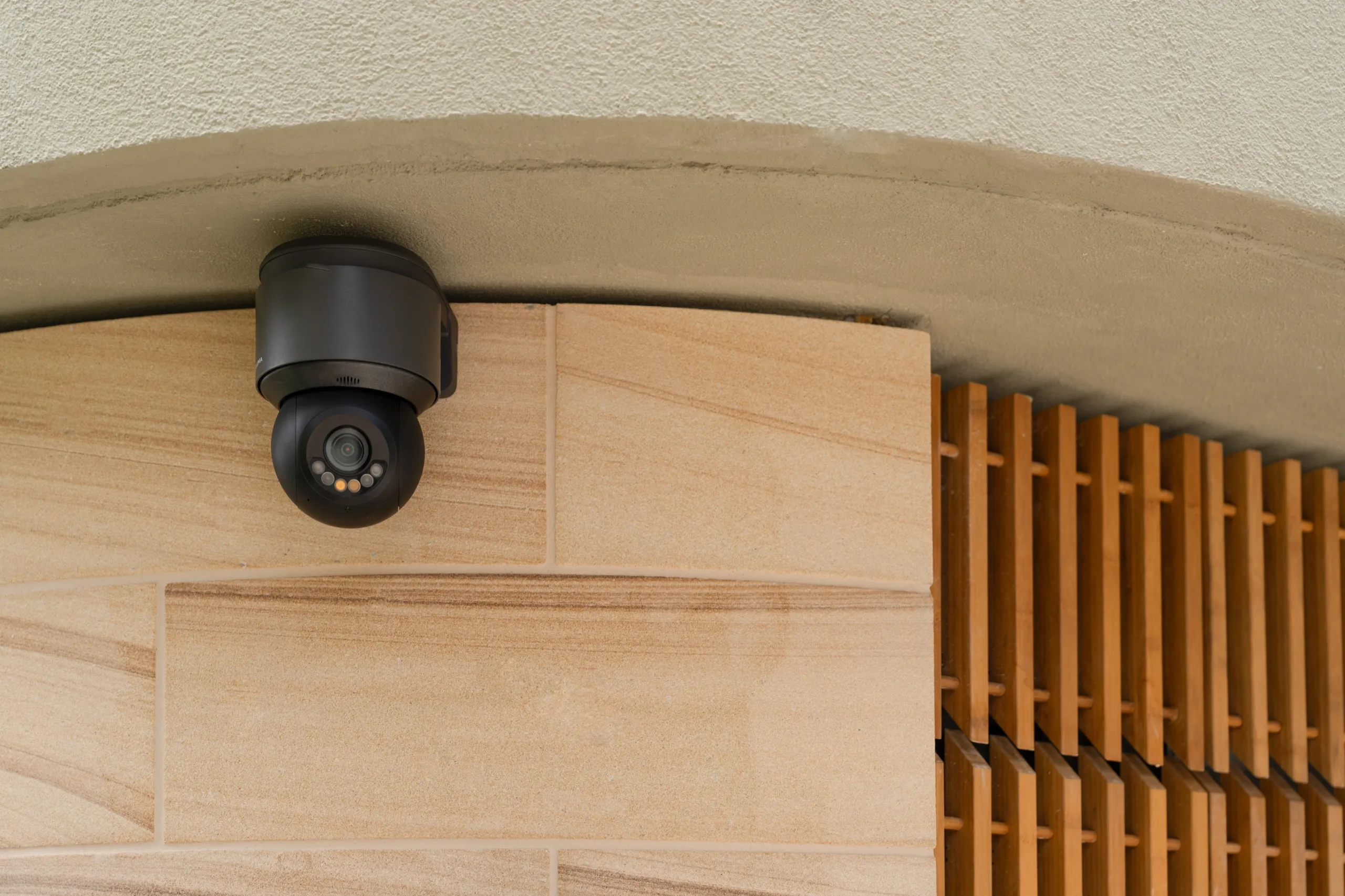 Dark gray outdoor dome security camera mounted on light stone cladding next to wood slat walling.
