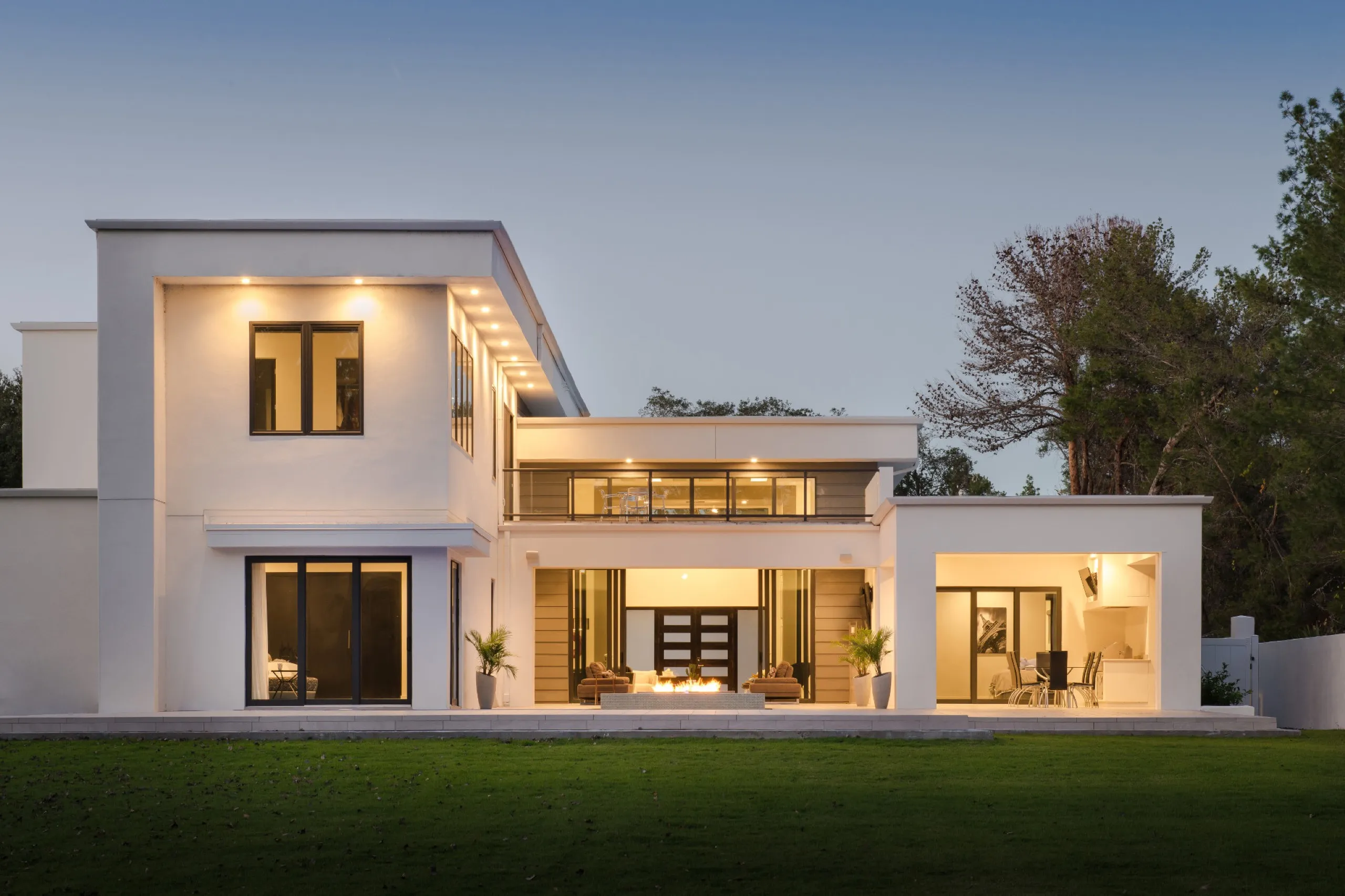 Modern white home at dusk with warm architectural lighting, a patio fire pit, and a broad green lawn.