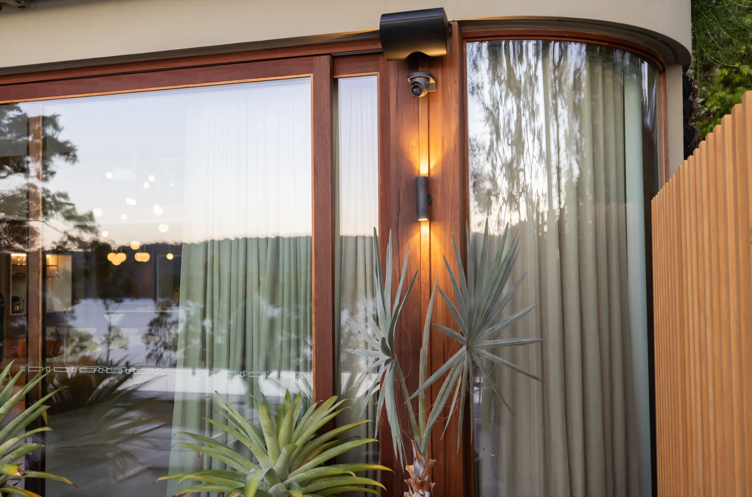 Black outdoor security camera and warm wall light on a curved wood-and-glass exterior with yucca plants.