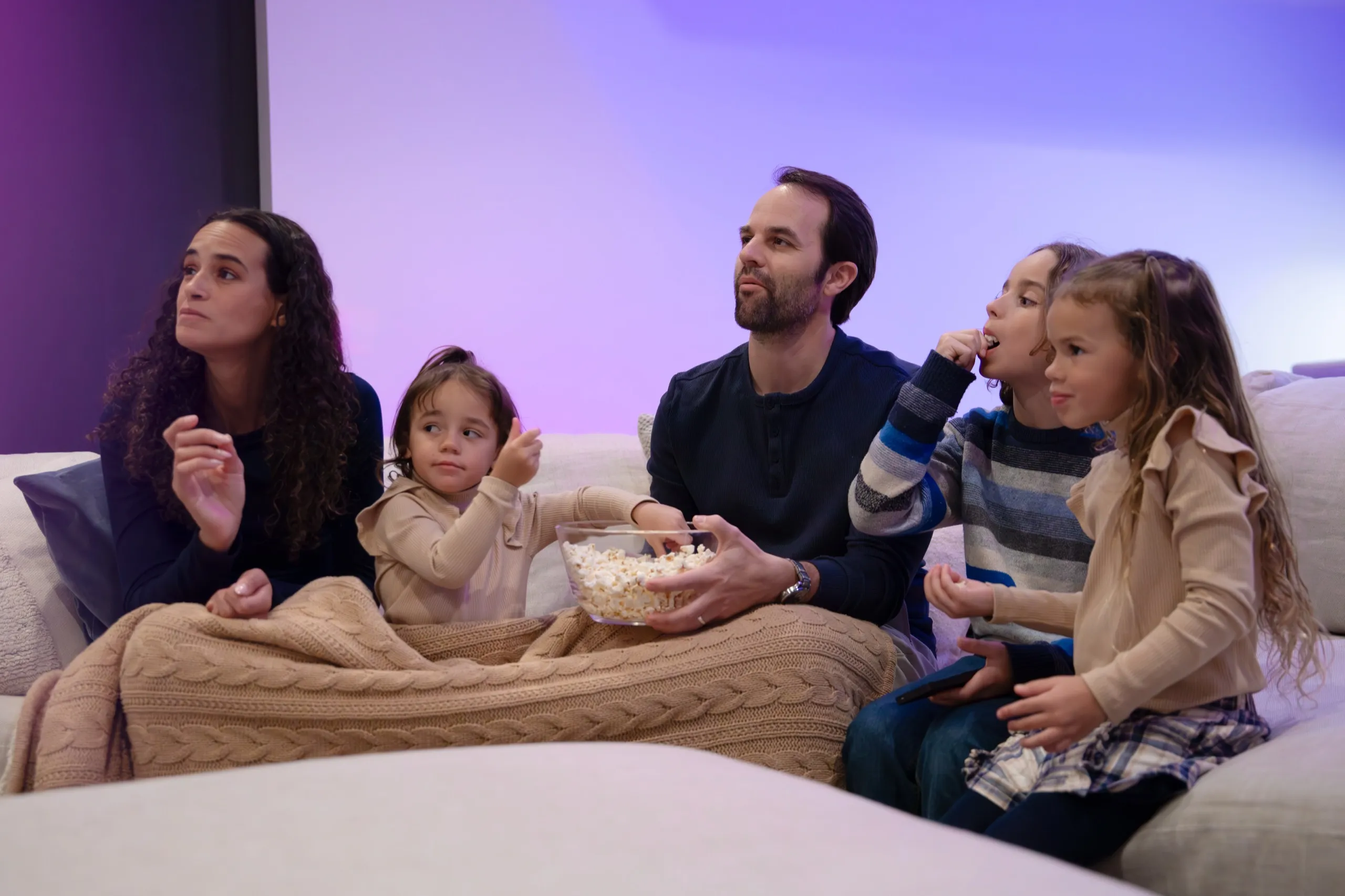 Family of five on a sofa with a bowl of popcorn watching a screen under purple ambient lighting.