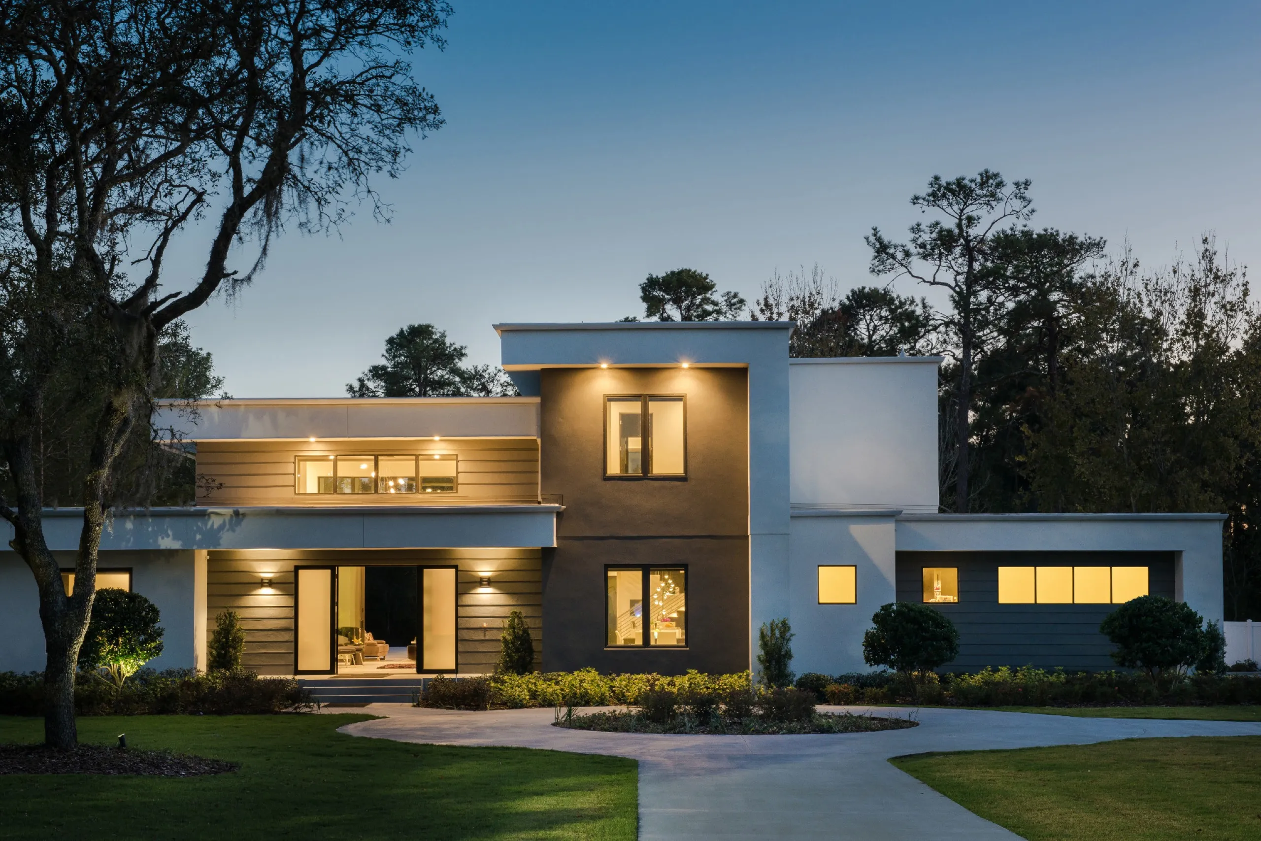 Twilight front view of a modern white and gray building with warm interior and exterior lighting along a curved driveway.