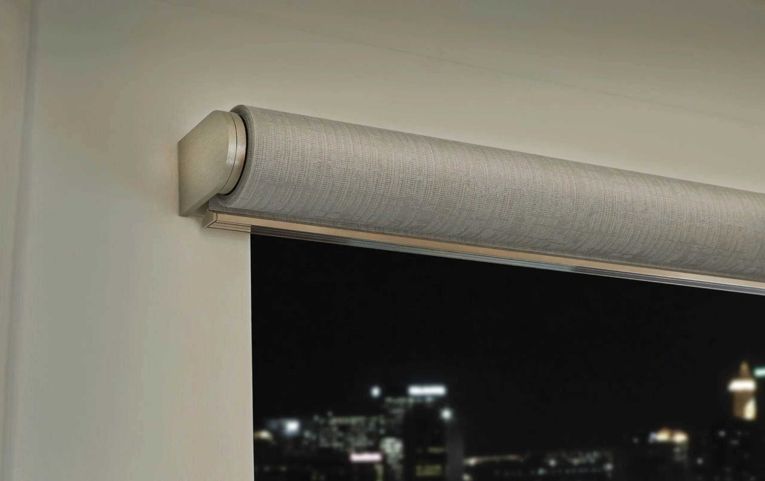 Close-up of a light gray roller shade tube with metal end cap above a night city skyline window.