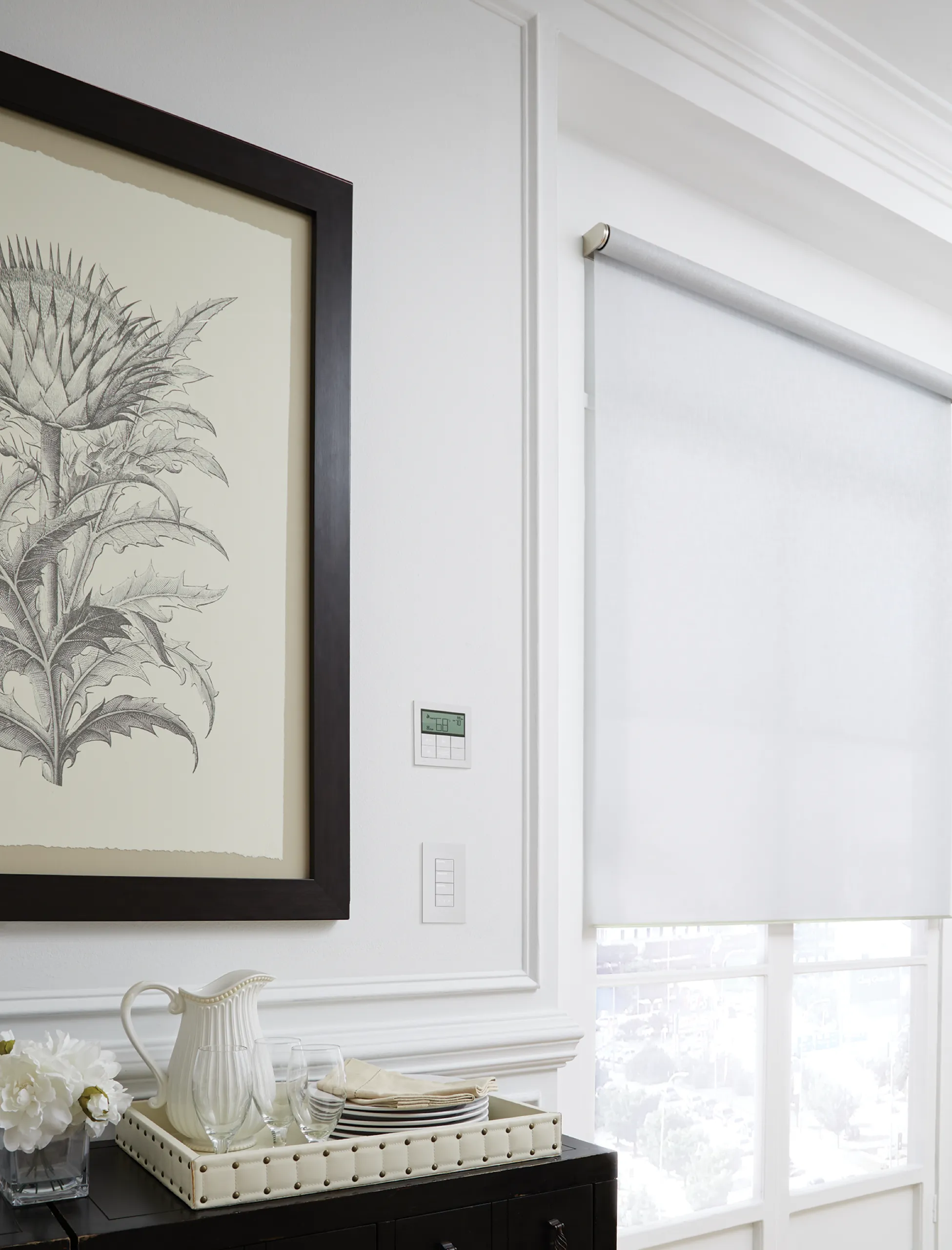 White wall with botanical print, digital thermostat, small keypad, and pale roller shade above a styled sideboard