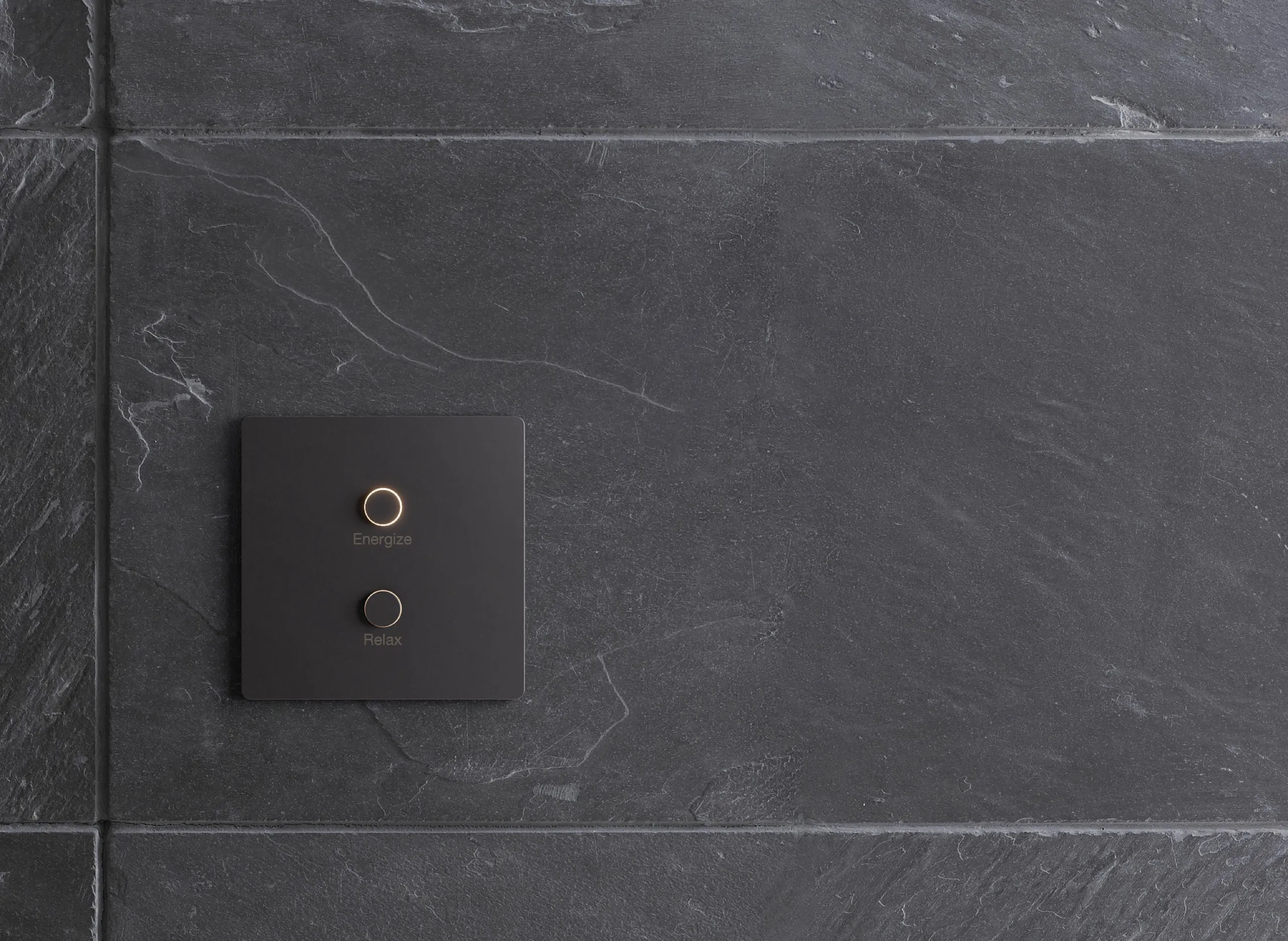 Dark charcoal two-button lighting keypad with the Energize ring lit gold, mounted on gray slate tile.