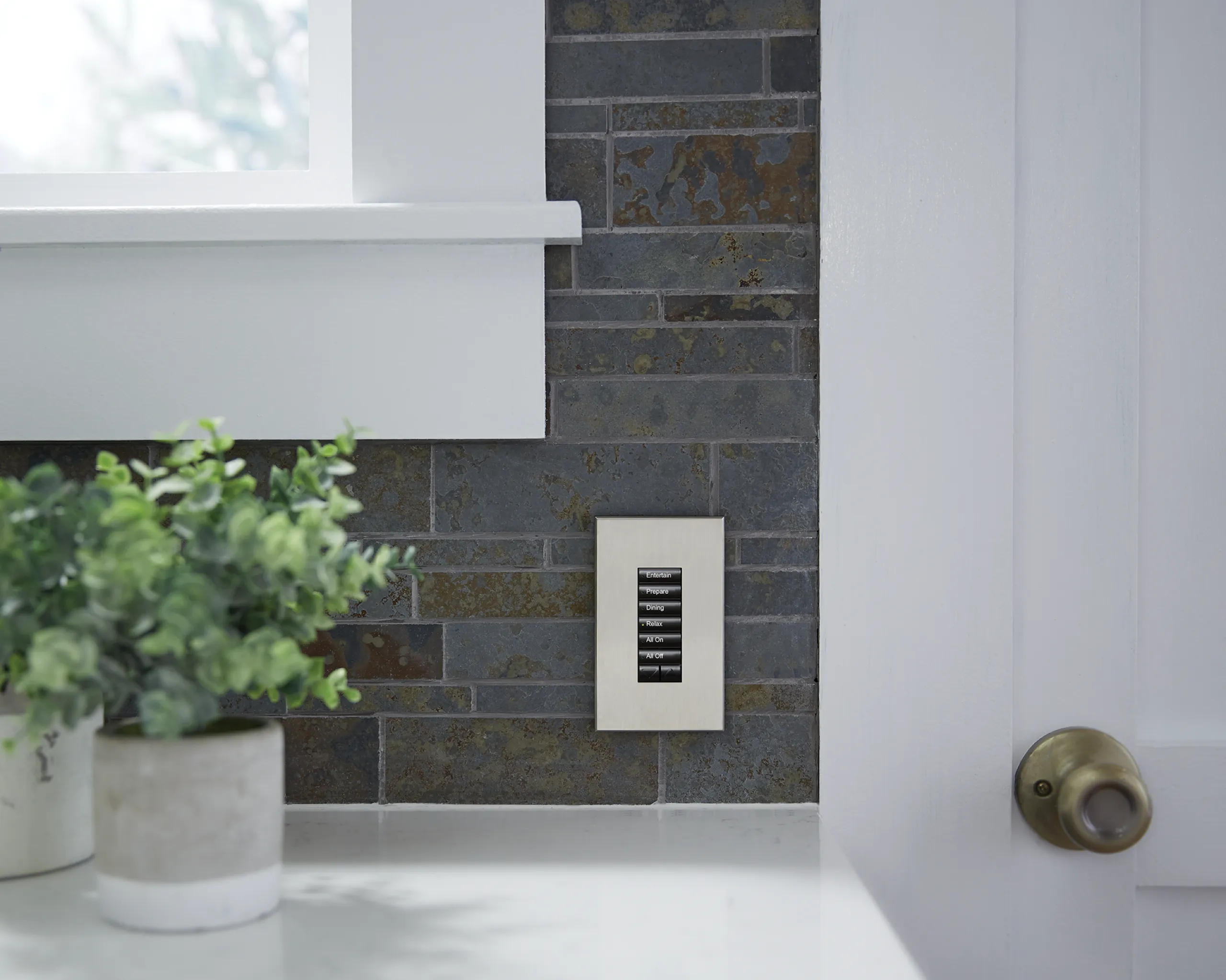 Silver Lutron seeTouch keypad with labeled scene buttons mounted on a dark stone kitchen backsplash.