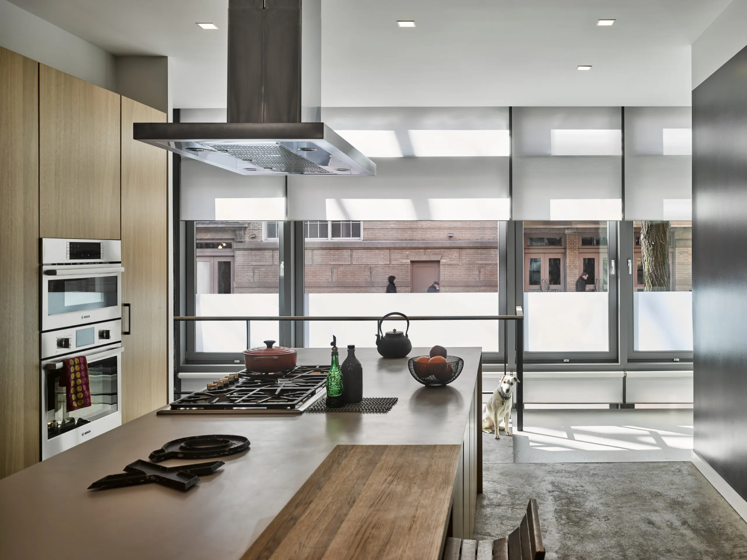 Modern loft kitchen with a concrete-floor island, stainless appliances, and white motorized shades on city windows.