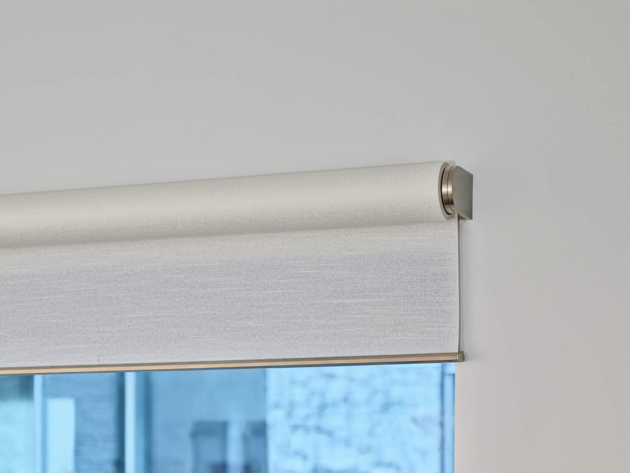 Close-up of a brushed-metal Lutron roller shade bracket and textured light fabric on a white wall.
