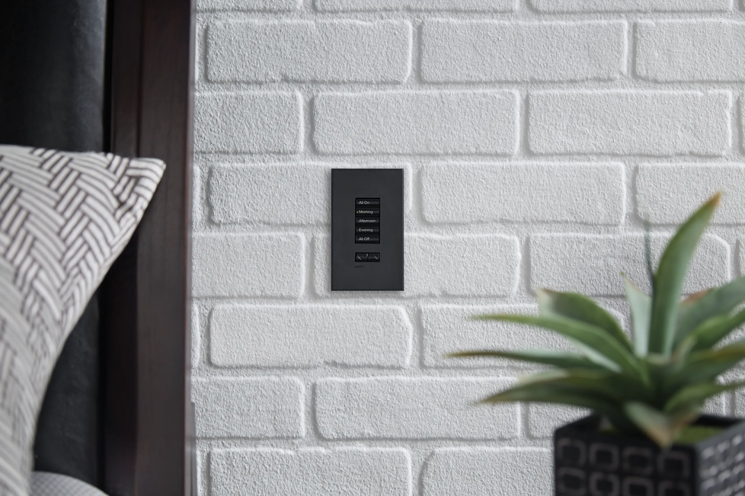 Black five-button scene keypad with All On through All Off labels mounted on white painted brick beside a bed