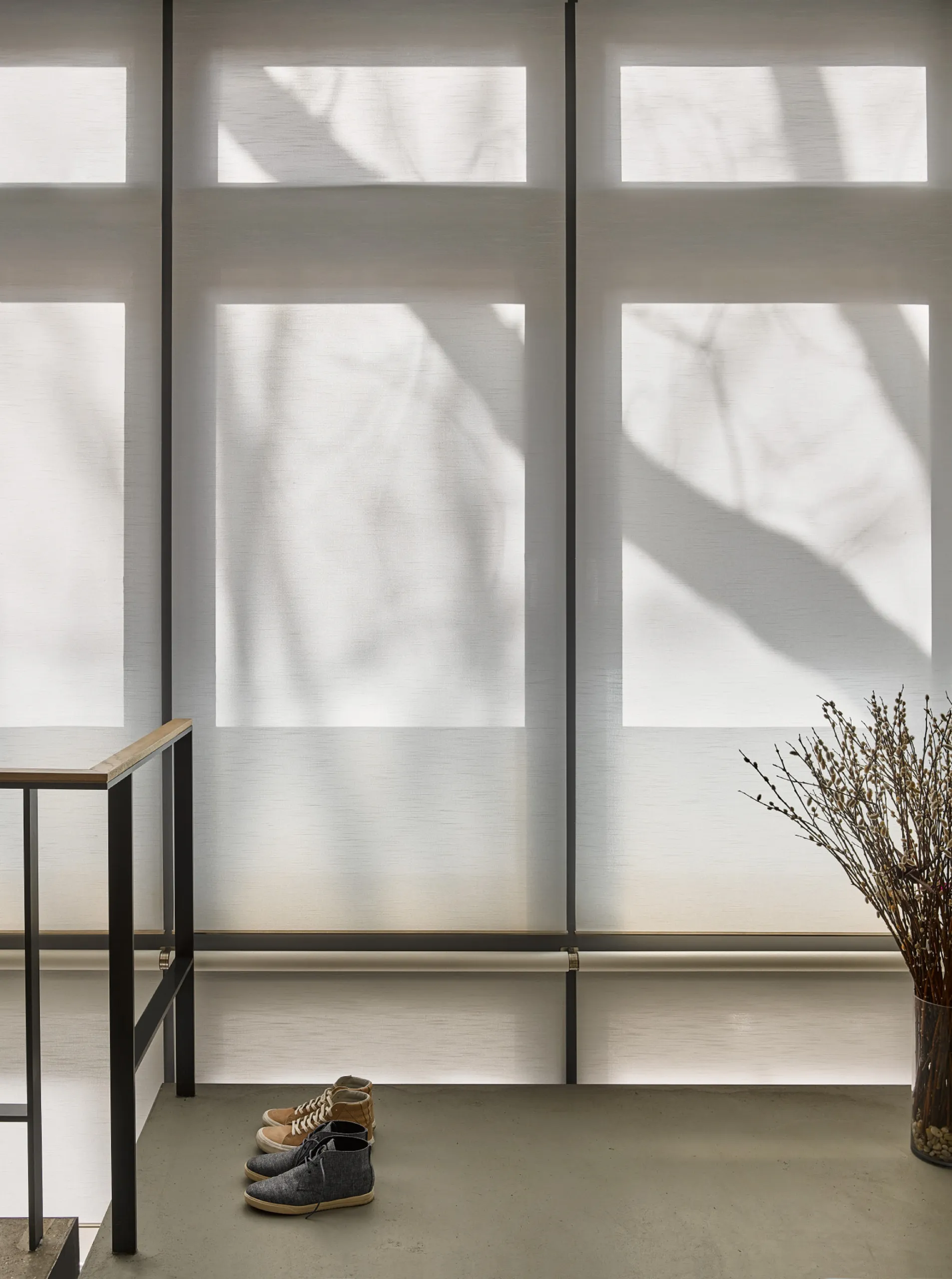 Tall loft windows with closed sheer white roller shades casting tree shadows beside a dark metal stair rail.