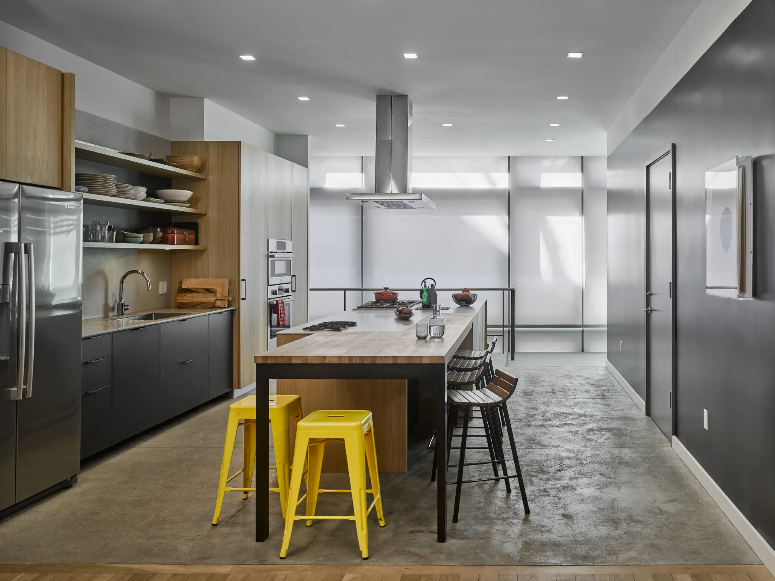 Industrial loft kitchen with a wooden island, yellow stools, and fully closed white roller shades.