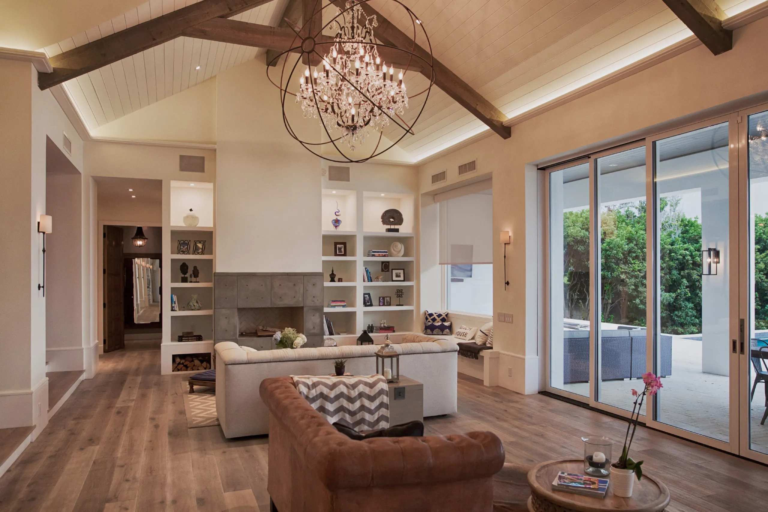 Open living room with vaulted beams, crystal orb chandelier, fireplace, and motorized shade beside patio doors.