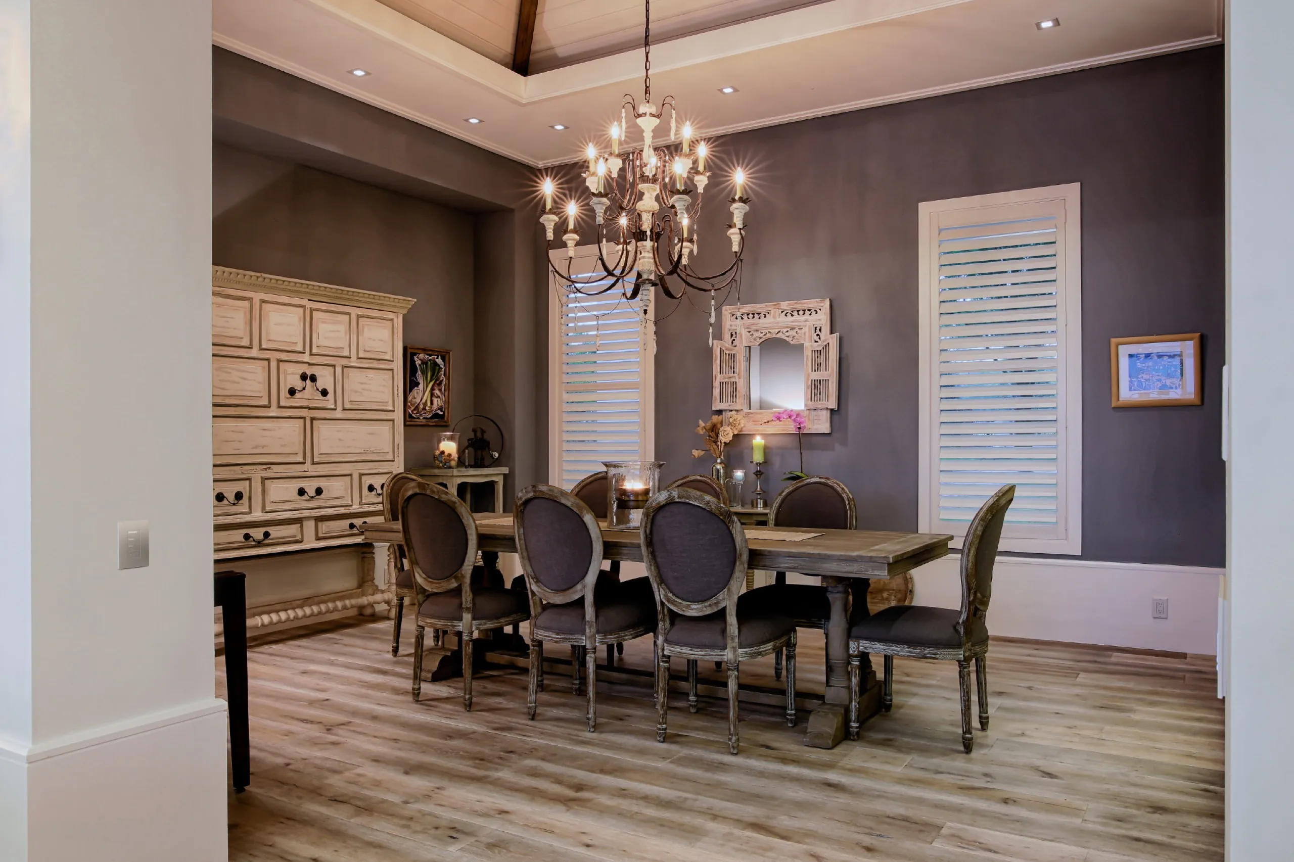 Traditional gray dining room with chandelier, plantation shutters, distressed hutch, and a small white wall keypad.
