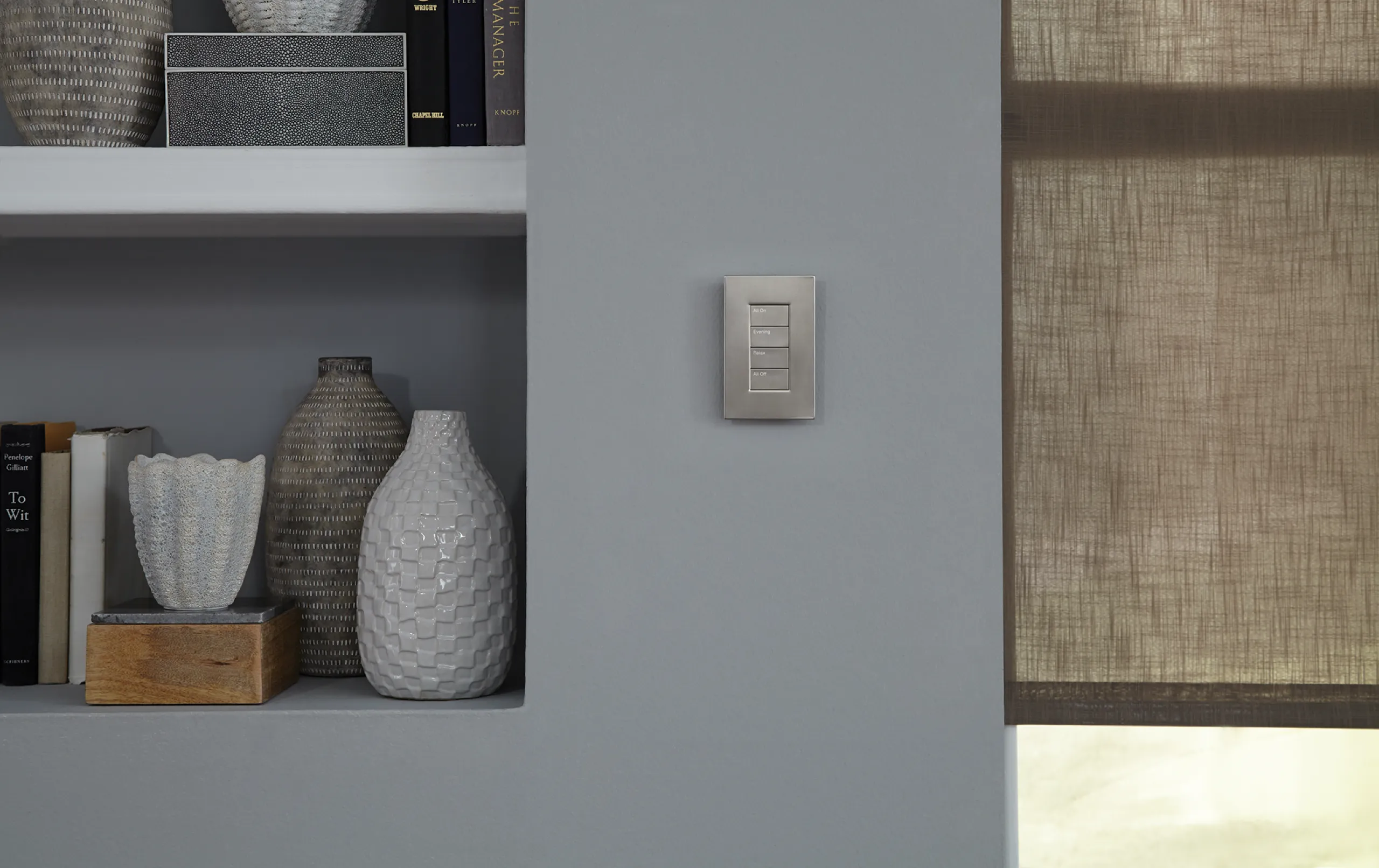 Satin nickel three-button wall keypad on a gray wall beside shelves of ceramics and a brown woven shade