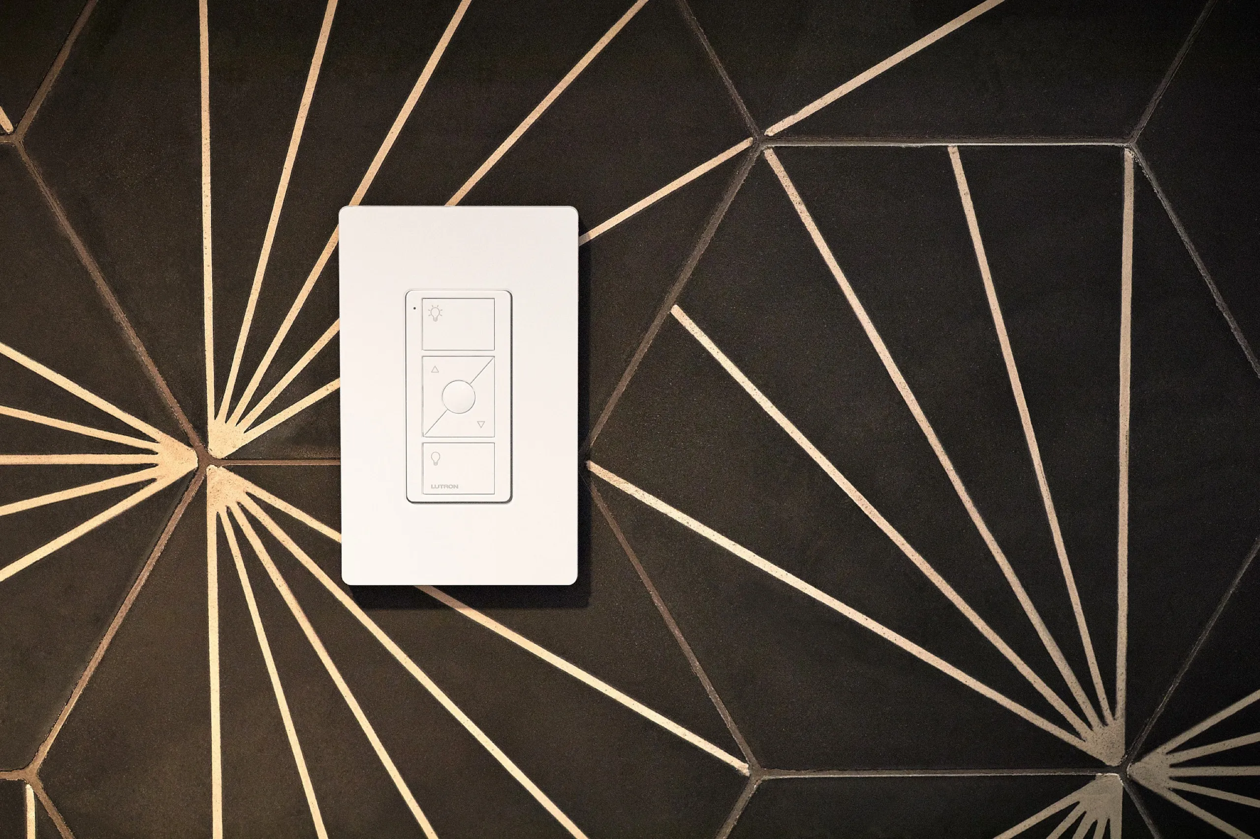 White Lutron Pico remote mounted on black hexagonal tiles with thin gold geometric lines.