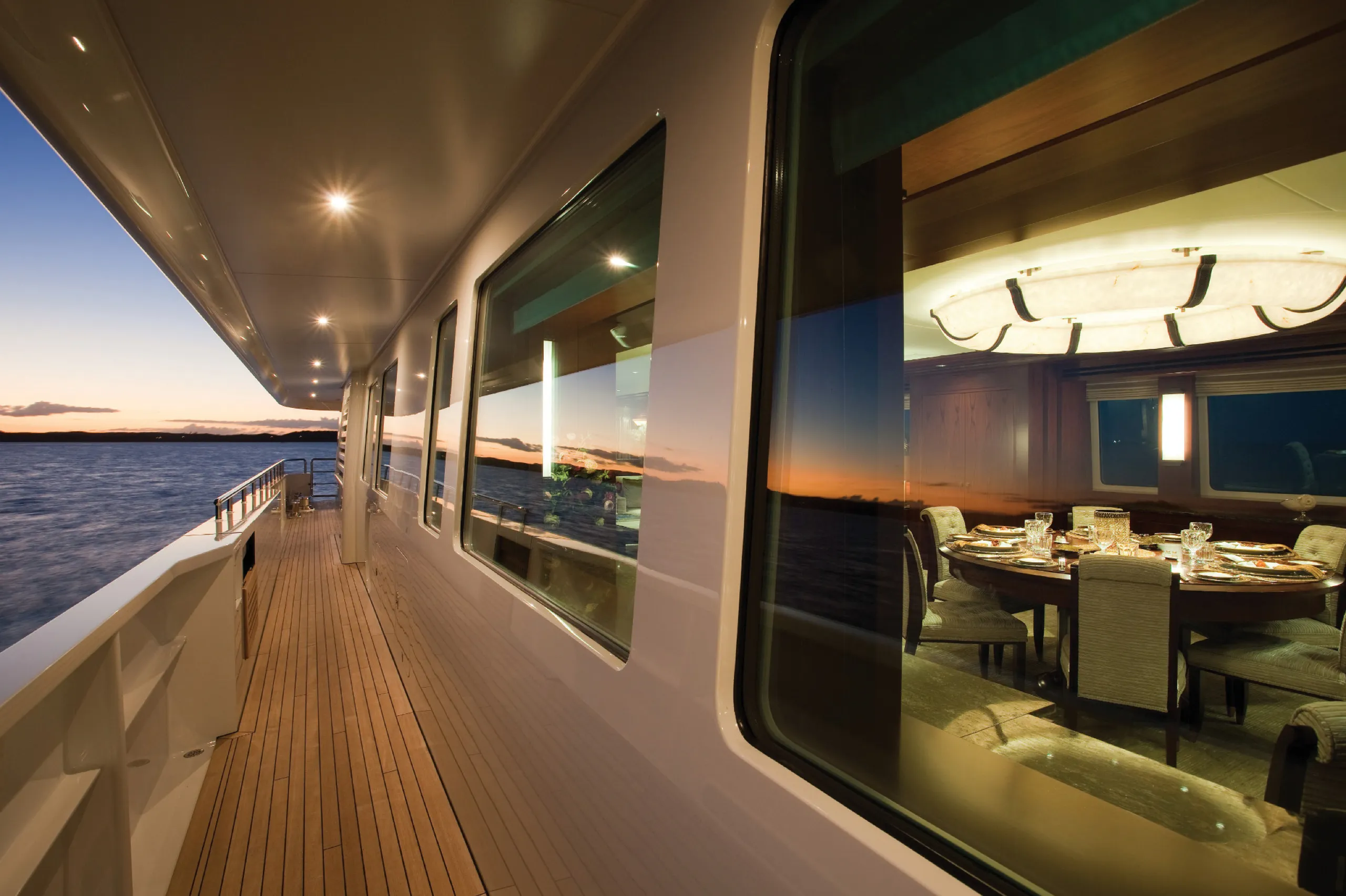 Long exterior walkway at sunset with teak decking and windows into a lit dining salon.