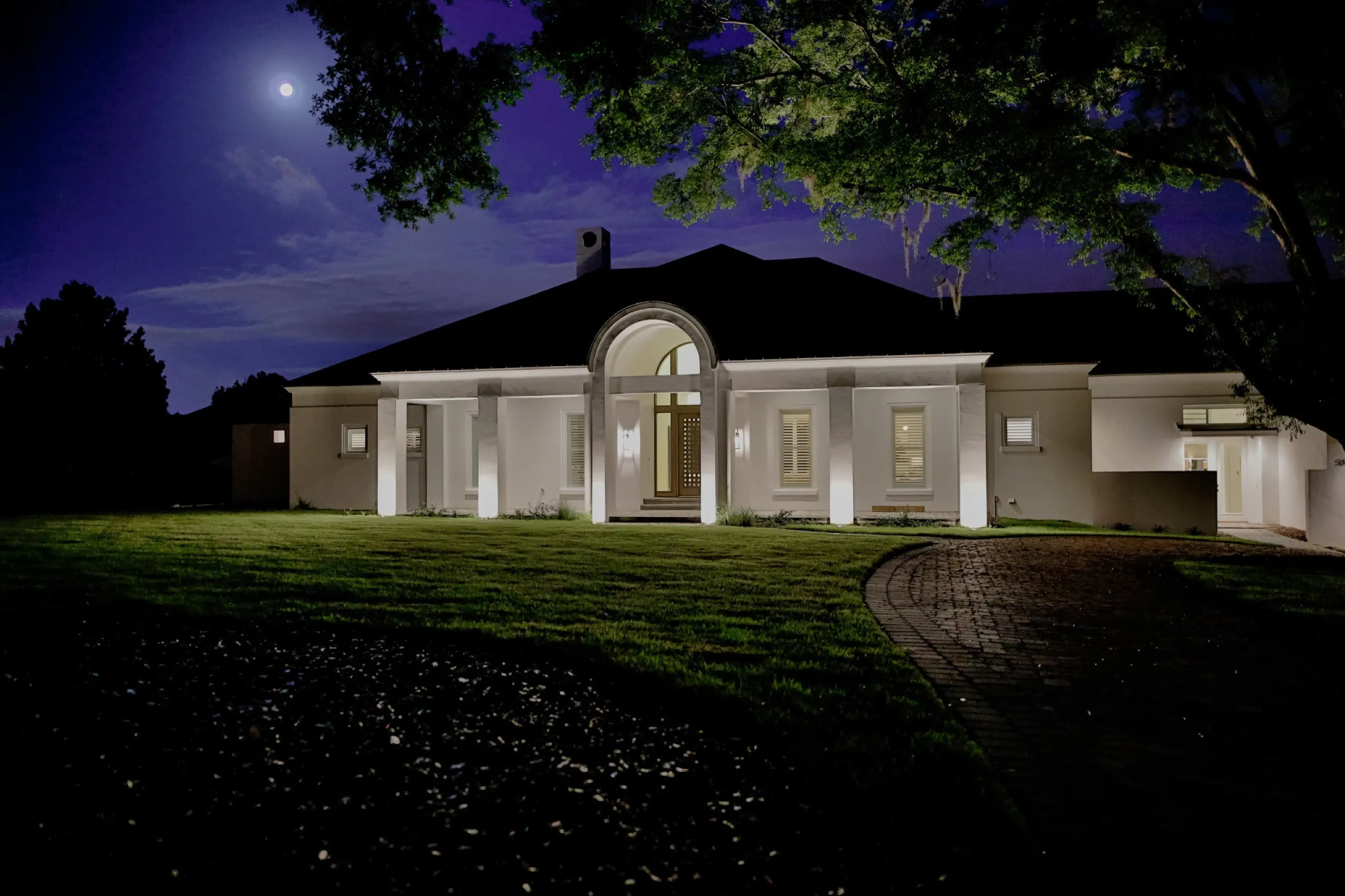 White columned home at night with warm facade uplighting, circular brick drive, and a moon in a deep blue sky.