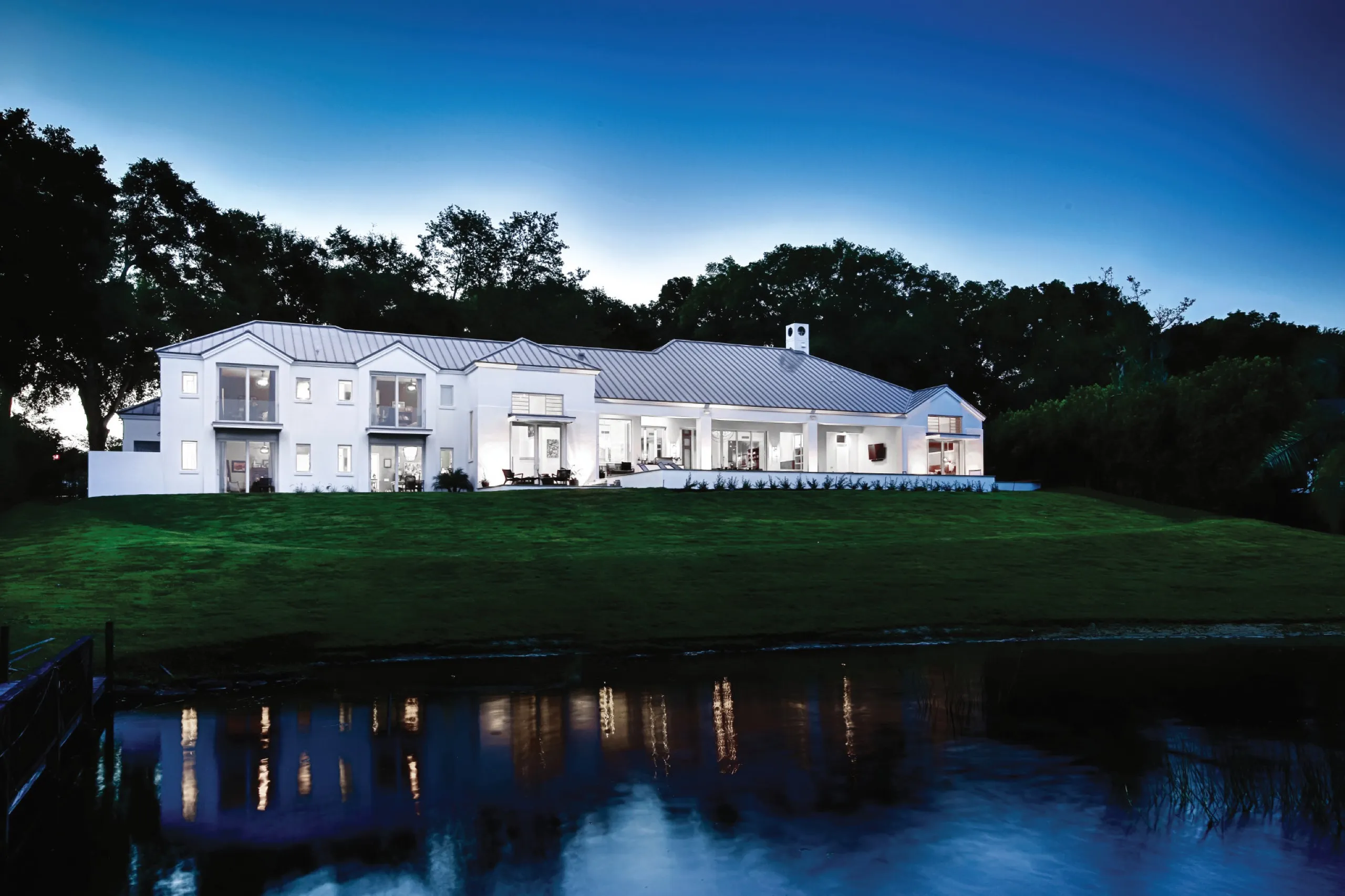 Large white house at twilight with architectural lighting reflected in a foreground pond.