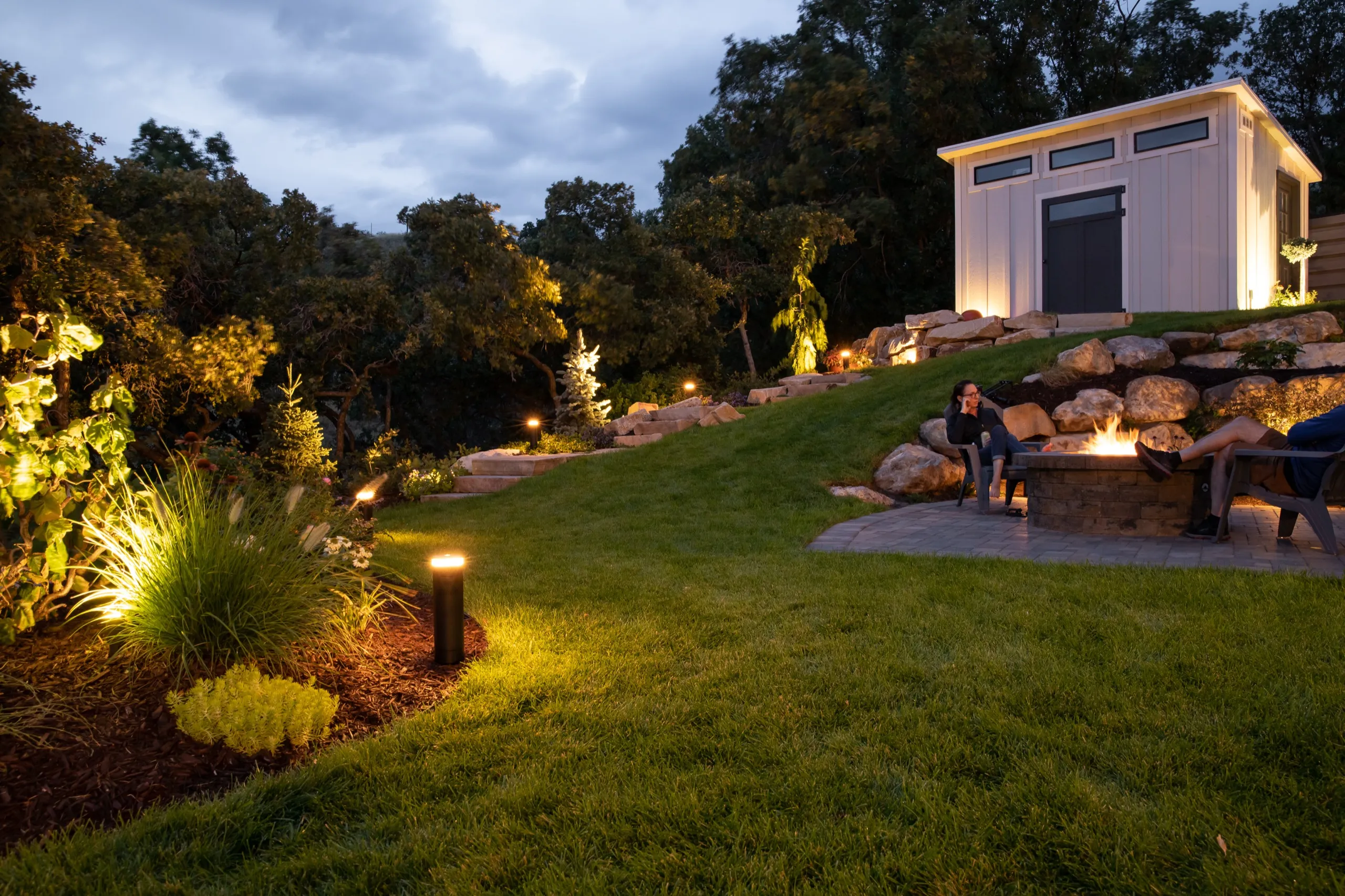 Evening backyard with path lights, a fire pit seating area, and a lit garden shed on a grassy hillside.