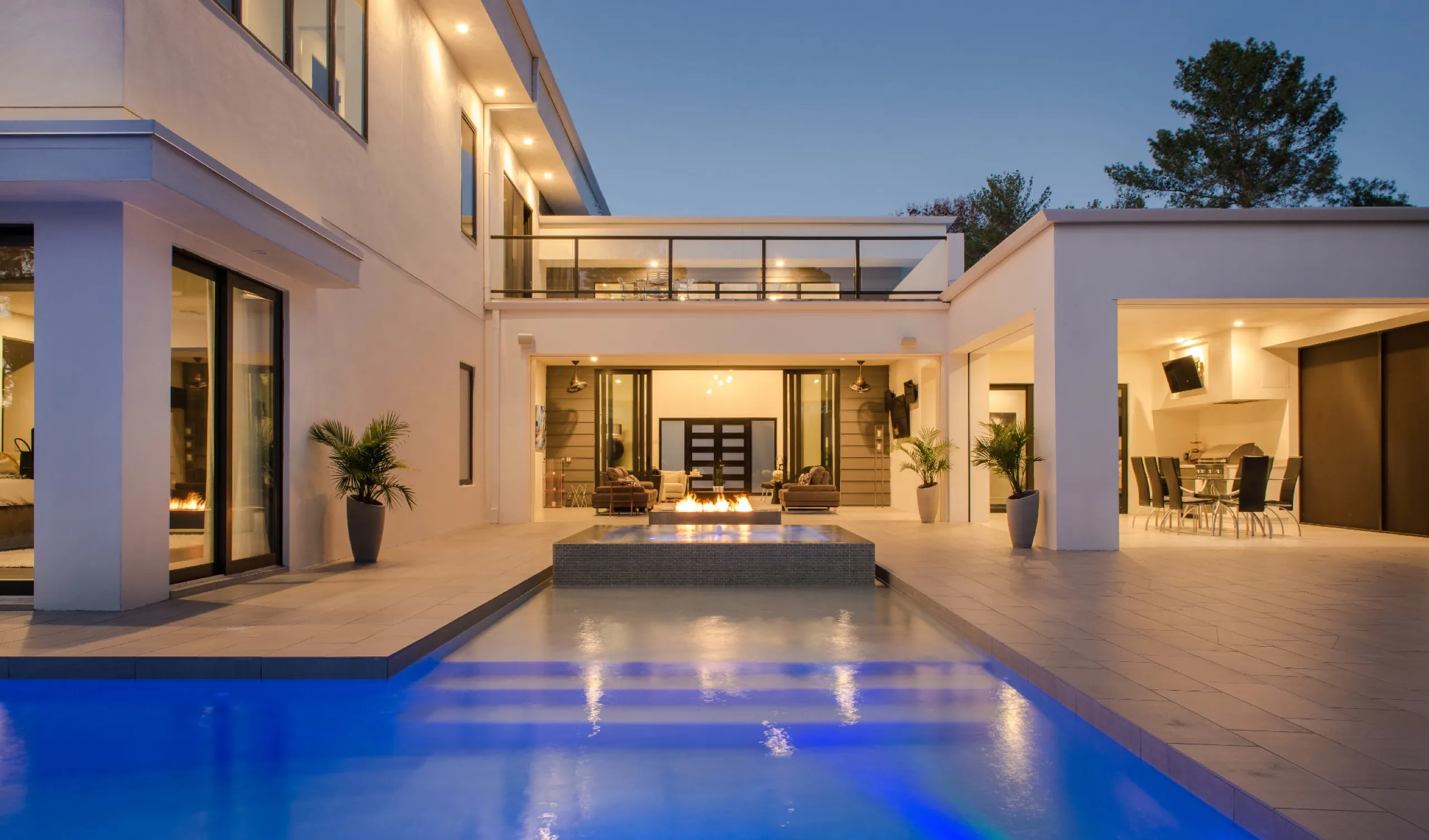 Luxury backyard at dusk with a glowing blue pool, spa fire feature, and covered outdoor dining pavilion.