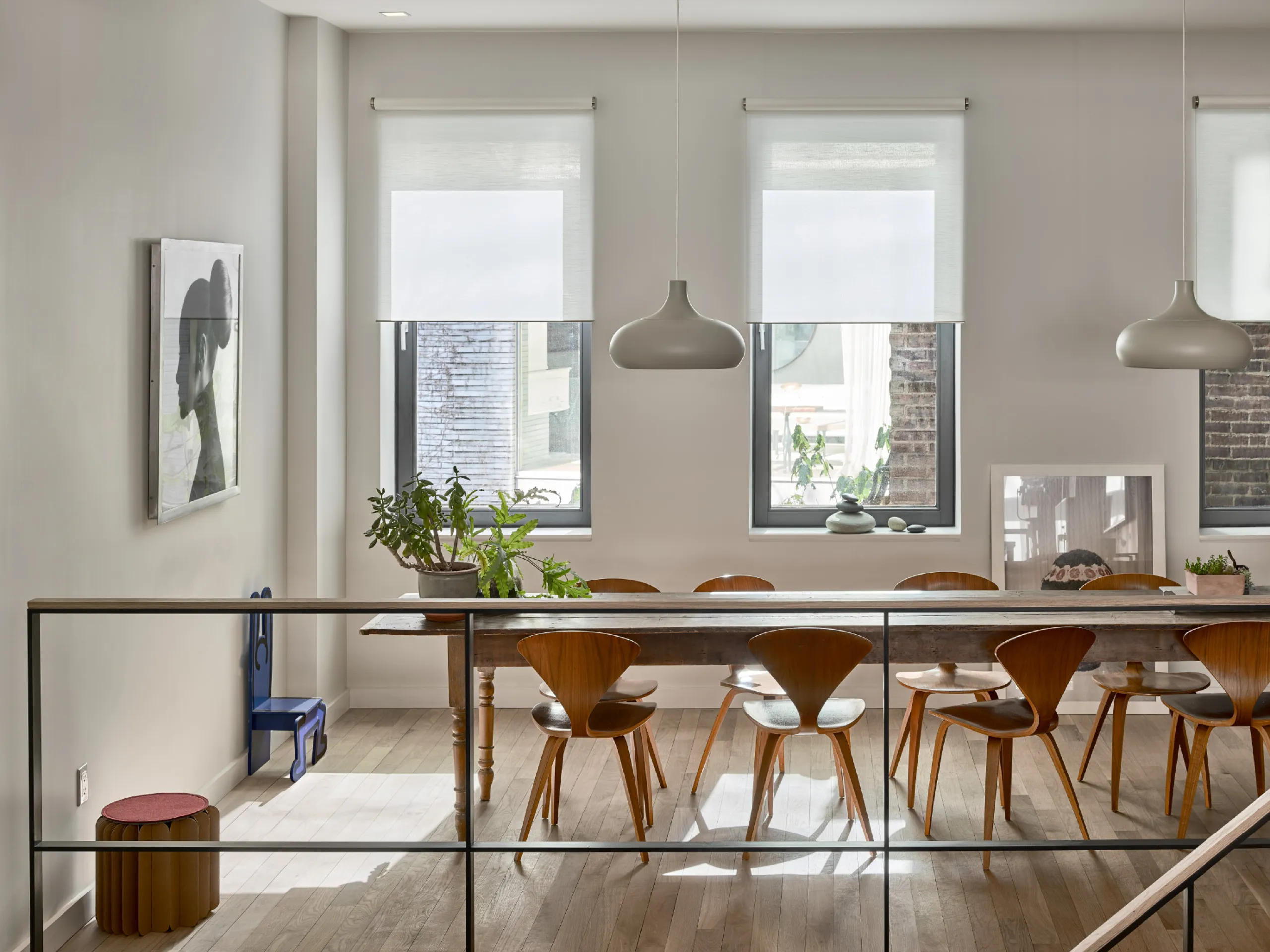 Loft dining area seen past a glass railing, with white roller shades on three tall windows and a long wood table