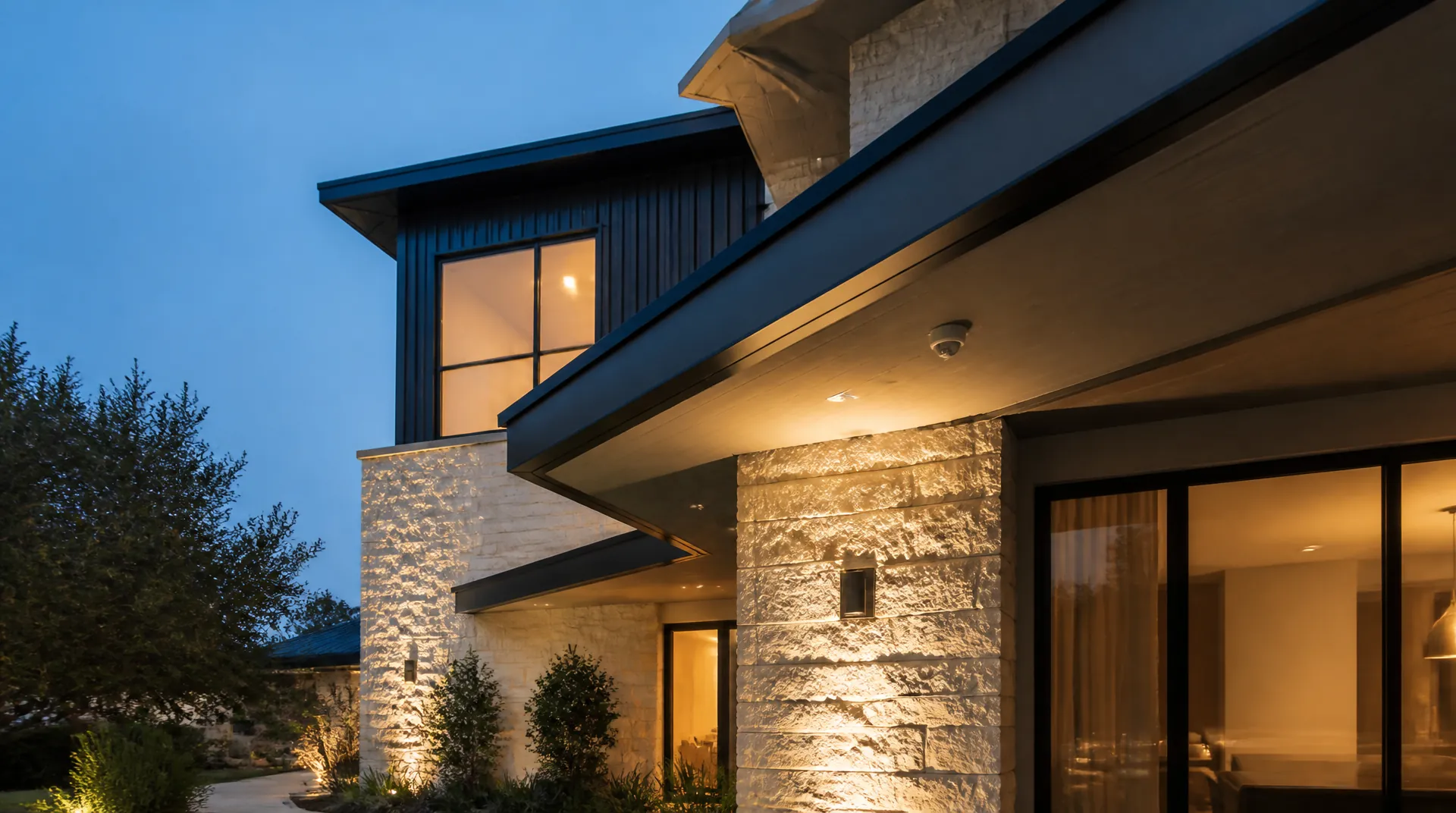 Dusk exterior of a limestone and dark-metal home with a small dome camera mounted flush under a deep eave above warm landscape lighting.
